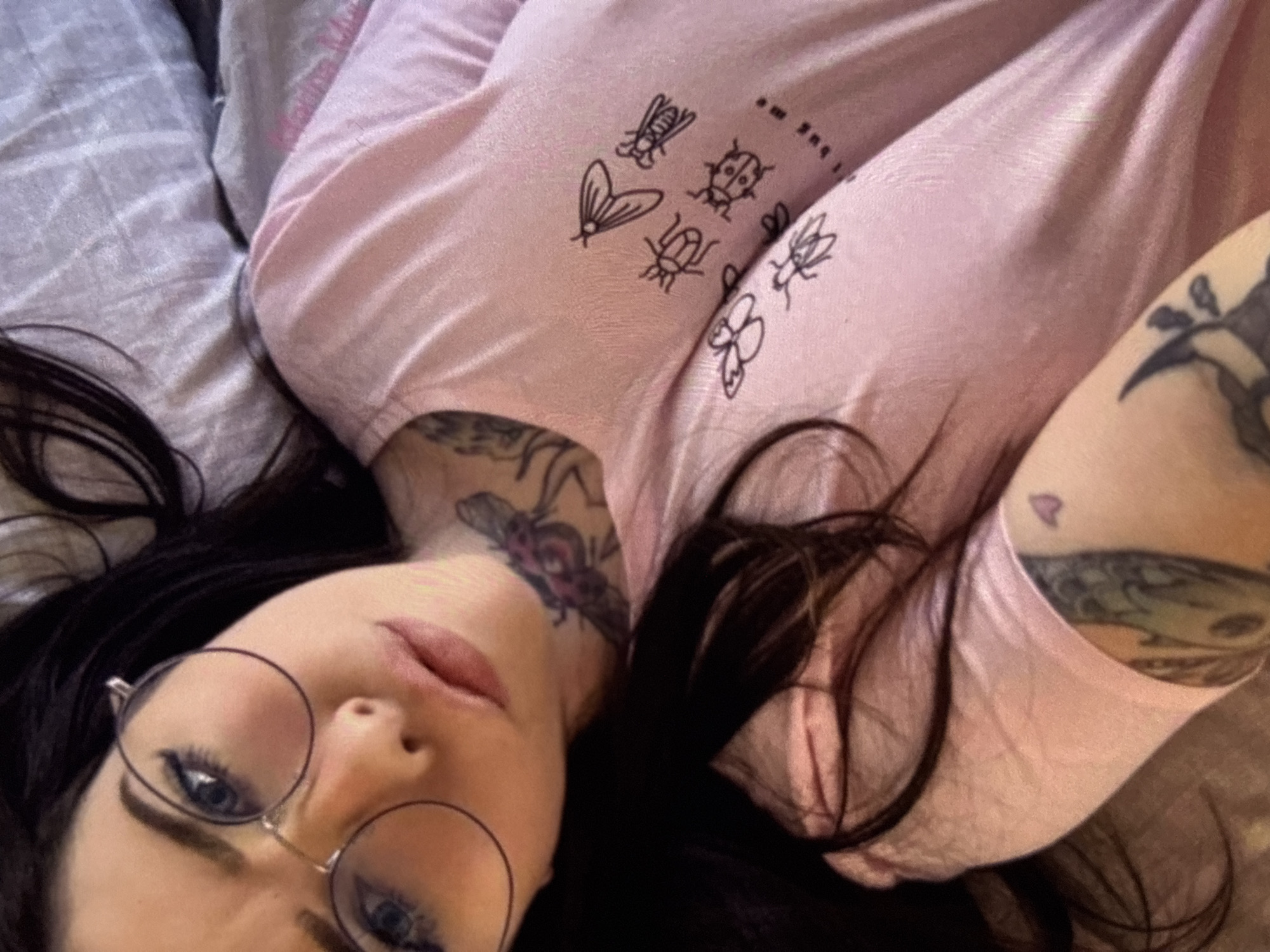 A close-up, overhead, portrait-style shot captures a young person with dark hair, wearing round glasses and a light pink t-shirt with a graphic print of various insects. The t-shirt has the text "Don't bug me" printed on it. The person's face is angled towards the camera, with their eyes looking upwards. A bee tattoo is visible on their neck. The background consists of a rumpled bed with a grey and purple patterned pillow. The scene appears to be indoors, likely in a bedroom.