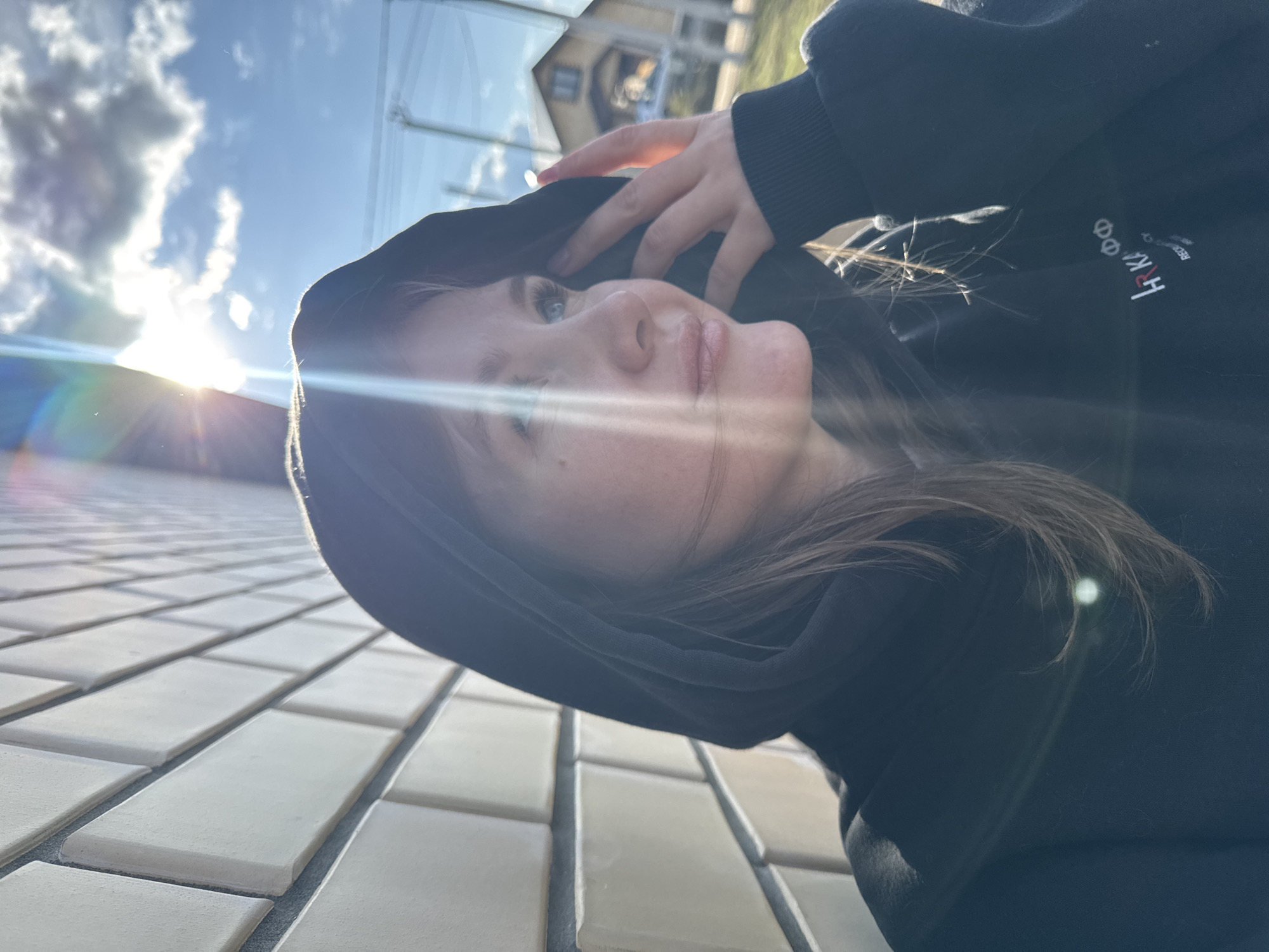 A young woman is the primary subject of this close-up photograph. She is wearing a black hoodie, with the hood up, and her hand is resting on top of the hood. The hoodie has a white logo on the upper right side. She has light skin, light eyes, and brown hair. The lighting is bright with sunlight shining strongly from the left side, creating lens flare and casting strong shadows.
The background shows a bright blue sky with white clouds and the partial silhouette of a house, likely located in a residential area, due to the presence of power lines. The foreground is a textured surface, possibly a wall made of light-colored rectangular blocks. The photo is taken at an angle, suggesting it may be a selfie.
The location is the city of Gornyi (Dvurechenskoe S/P), Russia.