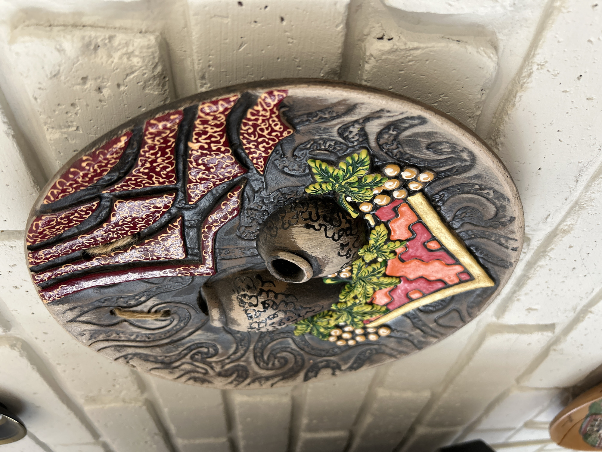 A circular, bas-relief ceramic plate is mounted on a white brick wall. The plate features a design with two clay pots, grapes, and stylized fabric or drapery. The pots are dark brown with black decorative patterns. Grapes and green leaves are depicted in the lower center, with colorful orange and red accents. The upper portion of the plate has a dark background with red and gold patterned elements, possibly representing fabric. The plate has a rope loop attached for hanging. The background is a textured white brick wall. The image is a close-up, with the plate occupying most of the frame. No people are visible in the image.