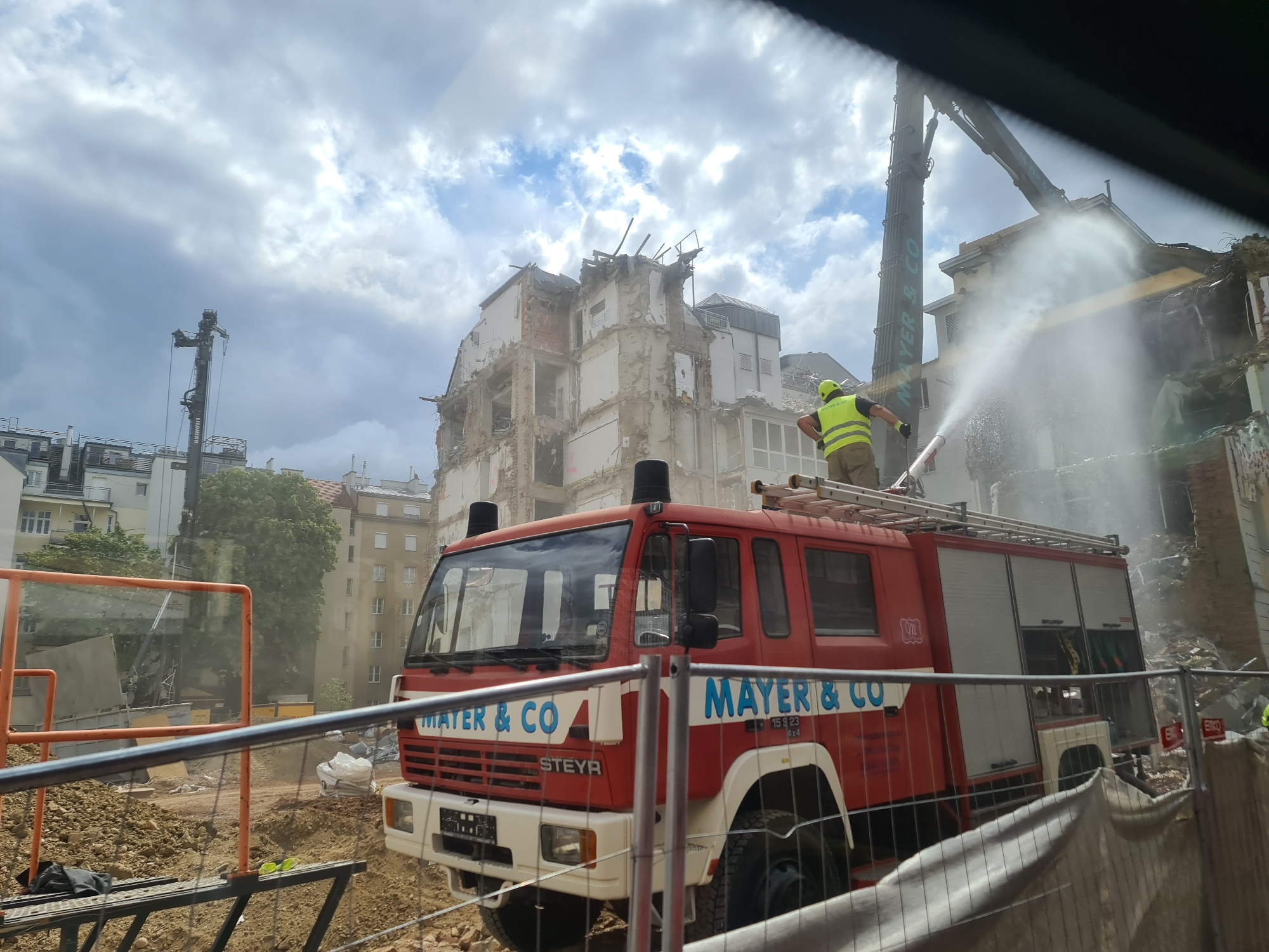 In Vienna, Austria, a demolition site is captured under a cloudy sky. A large building is in the process of being dismantled, with its upper floors severely damaged and exposed. A long-reach demolition excavator, bearing the "MAYR & CO" logo, is actively working on the structure. In the foreground, a red fire truck with "MAYER & CO" written on its side is parked. A worker, dressed in a yellow vest and hard hat, stands on top of the fire truck, operating a hose that is spraying a large plume of water, likely to control dust generated by the demolition. The ground around the site is covered in rubble and dirt, with construction fencing and barriers in place. The scene suggests an active demolition operation requiring dust suppression measures.