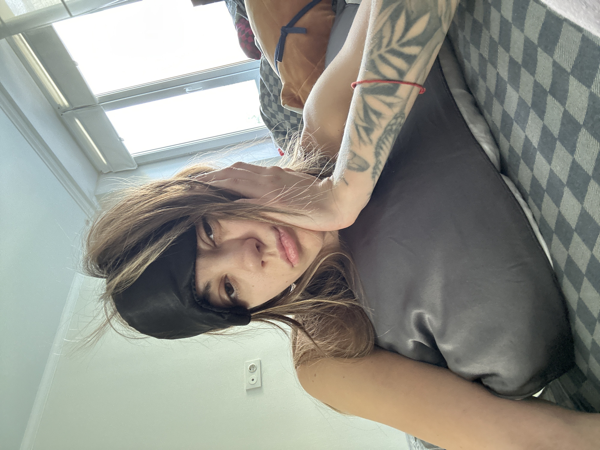 A young woman with a black sleep mask partially covering her eyes rests her head on a grey pillow, her face turned to the camera with a pout. Her brown hair is disheveled and appears to be sticking up in places, suggesting she has just woken up. Her left arm is propped on the pillow, supporting her head, and her right arm is extended across the pillow, revealing a tattoo of leafy branches on her forearm. A thin red string is tied around her wrist. She is wearing a sleeveless top. The background shows a window with blinds, allowing bright daylight to enter the room, and a wall with a light switch. The woman is lying on a bed with a checkered duvet. The scene appears to be a bedroom, likely in an apartment in Moscow, Russia.