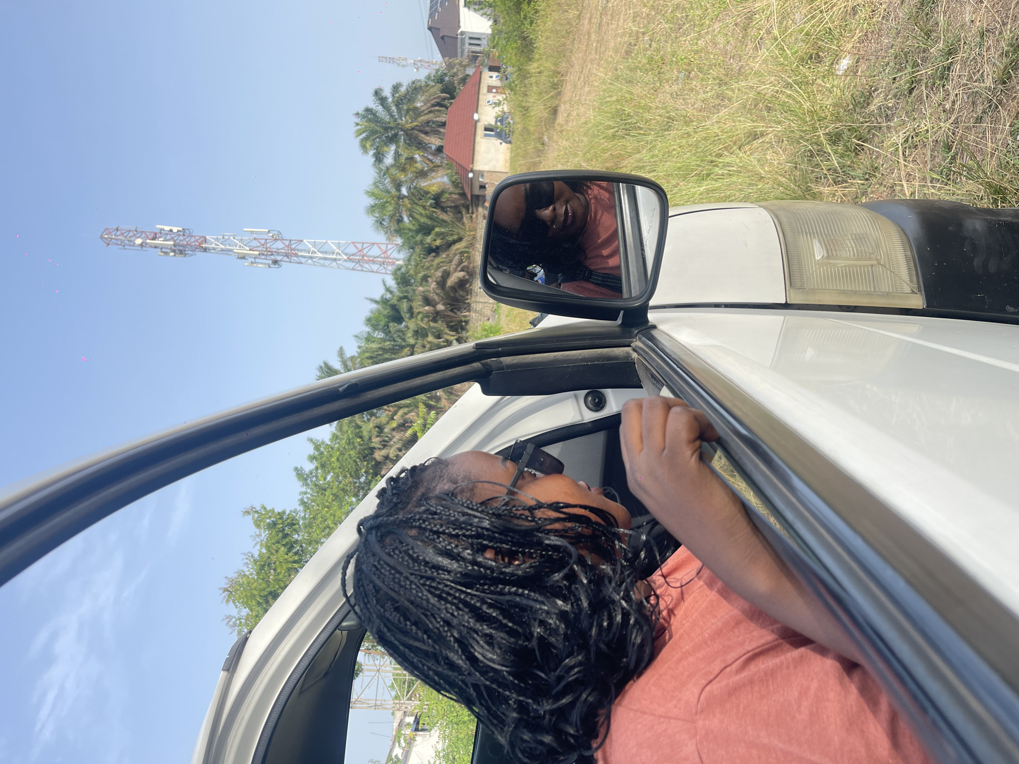 A medium shot of a person with braided hair leaning out the open door of a white vehicle. The person is wearing sunglasses and a salmon-colored top. In the side mirror, the reflection of the same person can be seen. Outside the vehicle, there is tall grass, palm trees, and a building with a red roof. A tall transmission tower with a white and red lattice structure is also visible against a blue sky. The city and location are unknown.