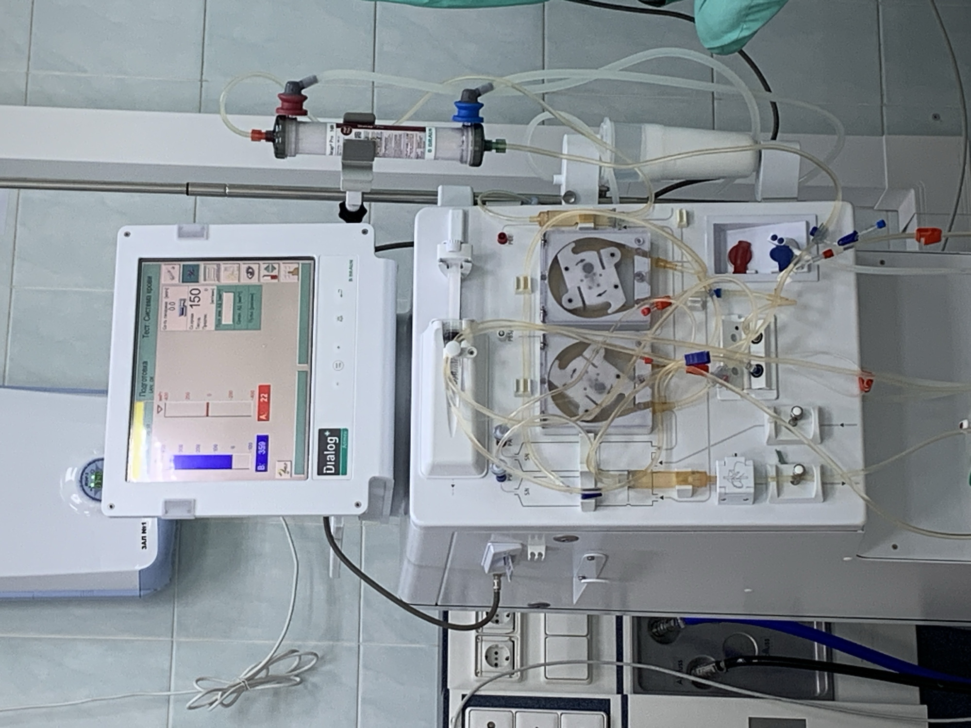 The image shows a Dialog+ hemodialysis machine with its components. The machine includes a screen displaying graphs and the word "Dialog+", along with various tubes, pumps, and connectors. A blood filter is positioned above the machine. The machine is mounted on a wall and set up in what appears to be a medical facility, likely a dialysis center. Based on the OCR text, the location is the city of Chaikovsky, Russia. Specifically, the equipment is likely located in hall number 1 (ЗАЛ №1) for hemodialysis.