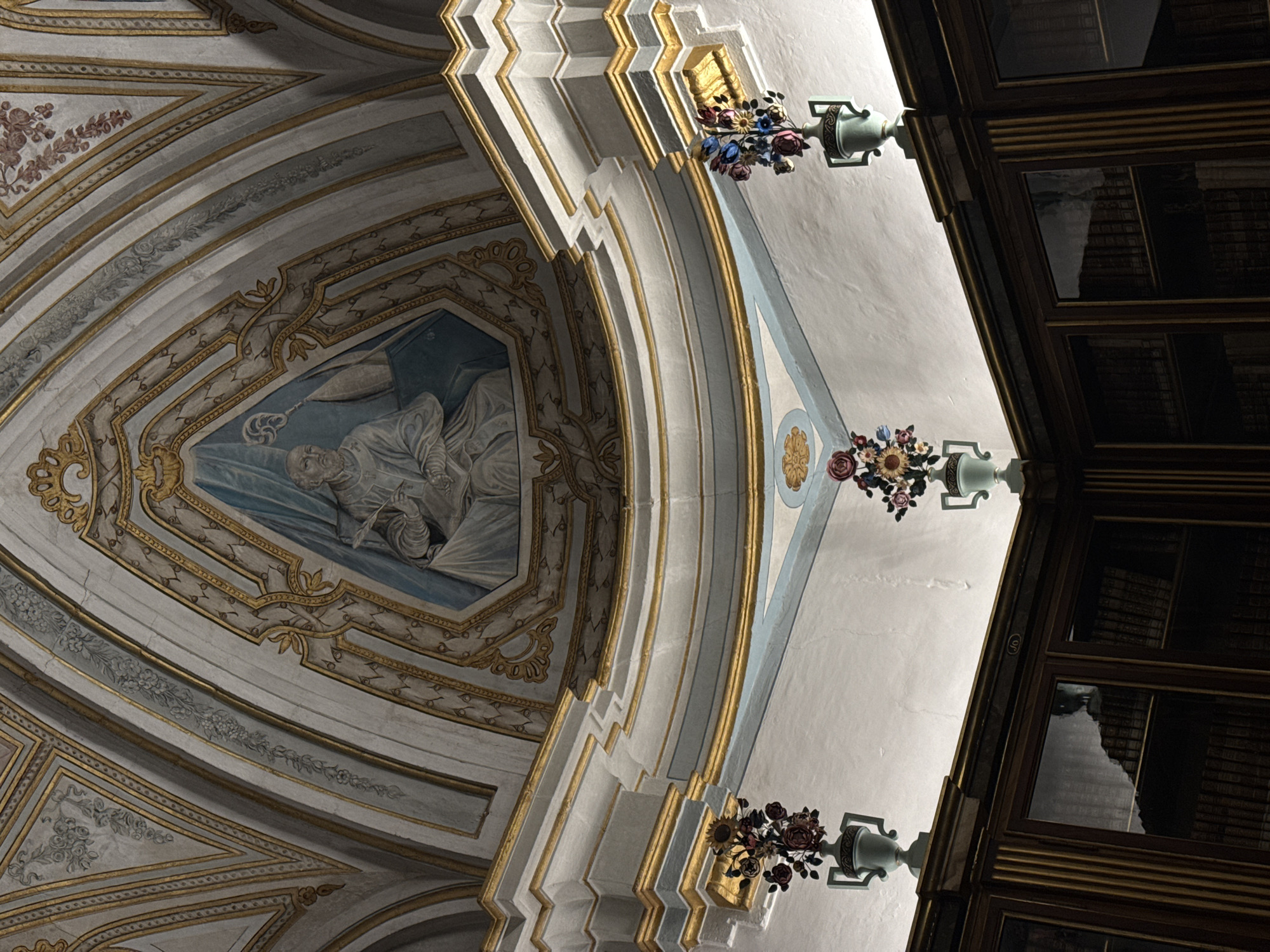 This media file depicts the interior of a grand, ornate building, possibly a church or a library, with a focus on the ceiling architecture and decorative elements. The ceiling is a complex structure of curves and recessed panels, adorned with gold trim and intricate painted designs. A significant fresco occupies a triangular recess in the ceiling, depicting a figure in repose, possibly in a religious or historical context. Surrounding the fresco are elaborate patterns and ornamental borders.
Attached to the ceiling are decorative light fixtures, resembling urns, from which cascades of artificial flowers hang. These floral arrangements add vibrant pops of color against the muted tones of the architecture. To the right of the image, a large, dark wooden structure with multiple glass panes dominates the frame. These panes reflect the interior of the building, hinting at the presence of bookshelves filled with books, suggesting a library setting. The lighting suggests it is daytime, with sunlight casting dramatic shadows, highlighting the architectural details and creating a sense of depth. The overall impression is one of historical grandeur and artistic richness, conveying a solemn and awe-inspiring atmosphere. No visible text is present in the image.