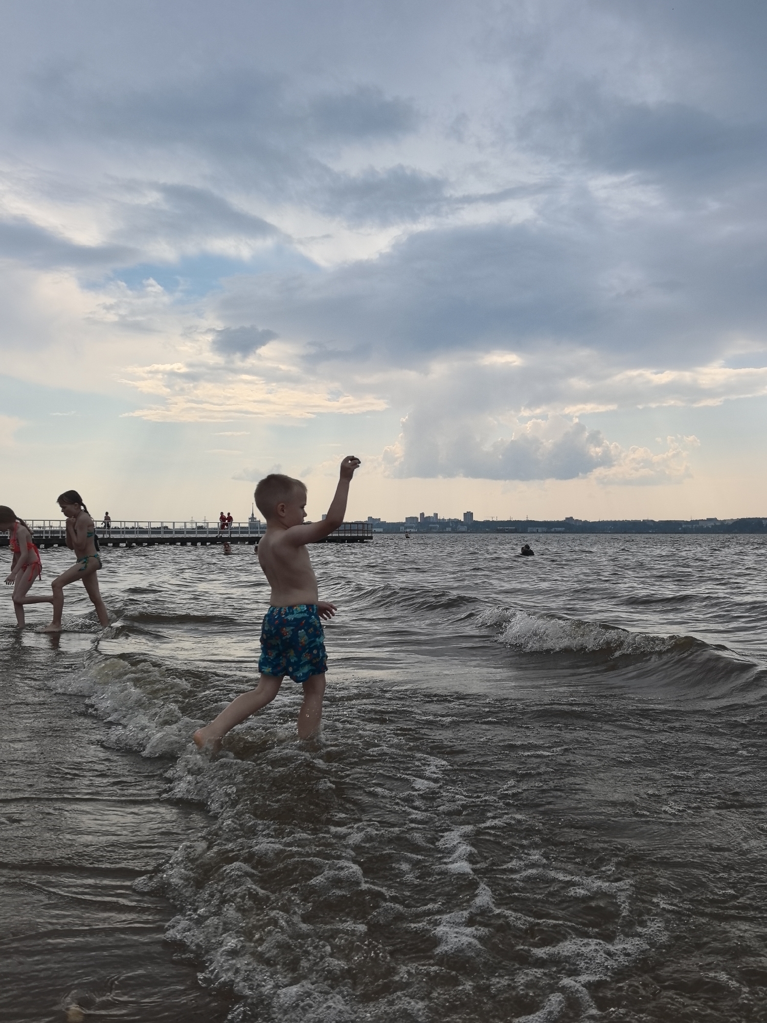 The image depicts a beach scene in Perm, Russia, with a cloudy sky overhead suggesting it might be late afternoon or early evening. The water is choppy, with small waves breaking near the shore, indicating a breezy or unsettled weather condition.
In the foreground, a young boy in patterned swim shorts is walking through the water, his right arm raised as if throwing something. To his left, two girls in swimwear are also wading in the shallows. One girl, in a bikini, is looking down at the water, while the other, in a patterned swimsuit, is walking forward.
In the background, a long pier extends into the water, with several people visible on it. Further in the distance, a city skyline can be seen on the horizon, providing a clear indication of the location. The overall mood seems to be one of casual summer recreation, with children enjoying a day at the beach despite the overcast sky. There is no visible text in the image.