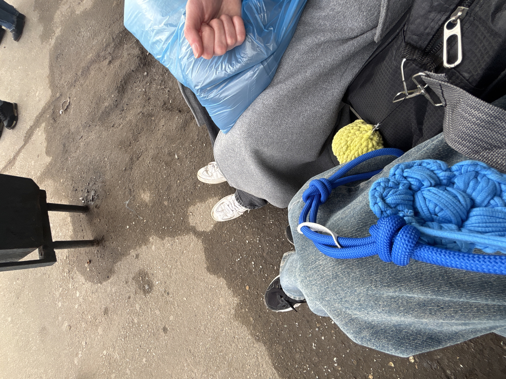 The image captures an overhead view of two individuals seated on what appears to be a public bench or seating area in Perm, Russia. The setting is an urban environment with a concrete ground surface.
The person on the left is wearing grey sweatpants and white sneakers, holding a crinkled blue plastic bag. The person on the right is wearing blue jeans and black sneakers. A blue, knotted, crocheted bag rests on their lap. A yellow crocheted ball is attached to a black backpack which is between the two people.
To the left, a black metal chair or bench leg is visible, indicating additional seating. The ground shows signs of wear and contains various small debris.