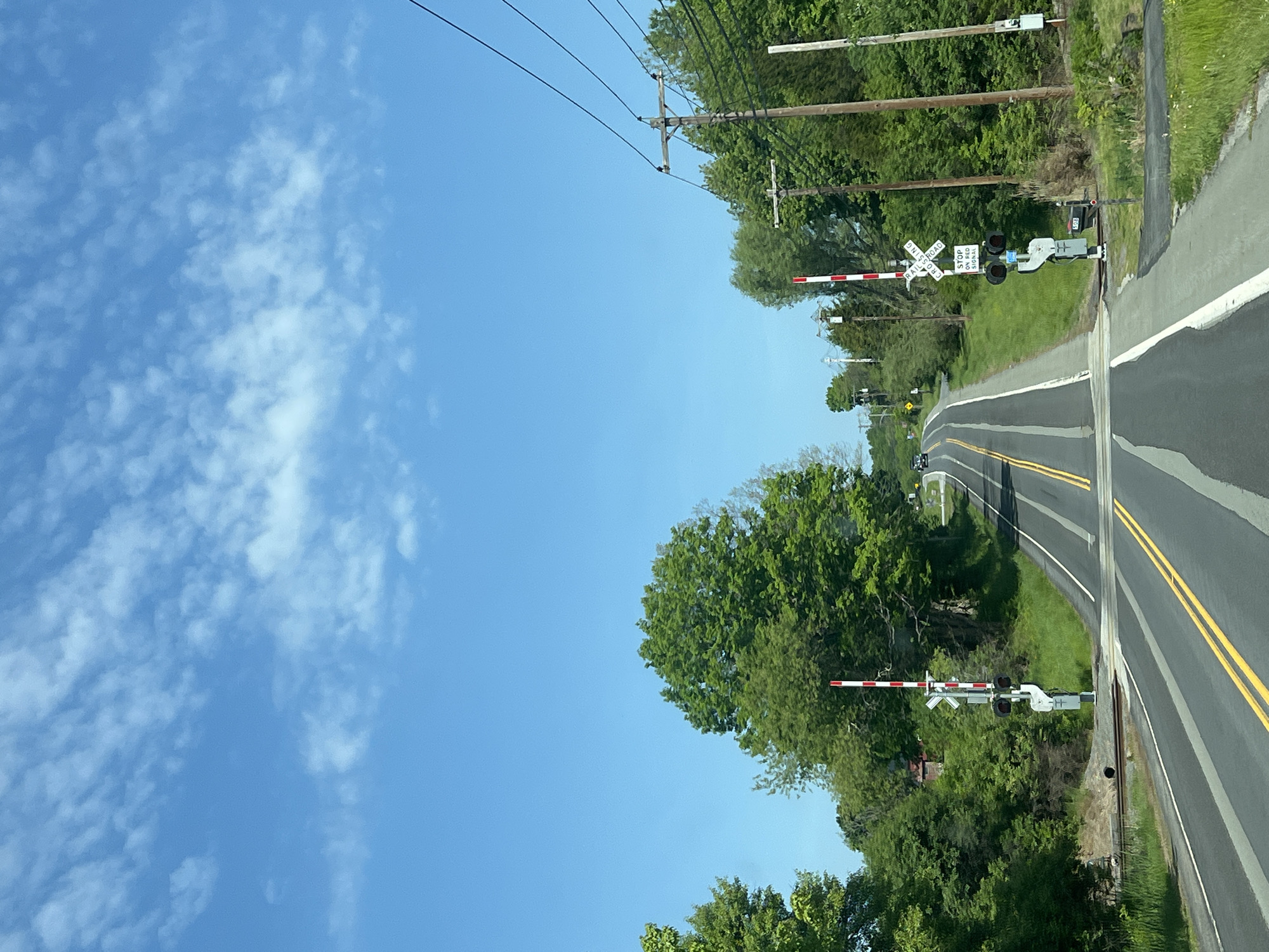 The image depicts a road in the Town of Chester, United States of America, featuring a railroad crossing with gates. The road is paved with double yellow lines. Trees line both sides of the road. Overhead power lines run along one side of the road. The sky is blue with some clouds.