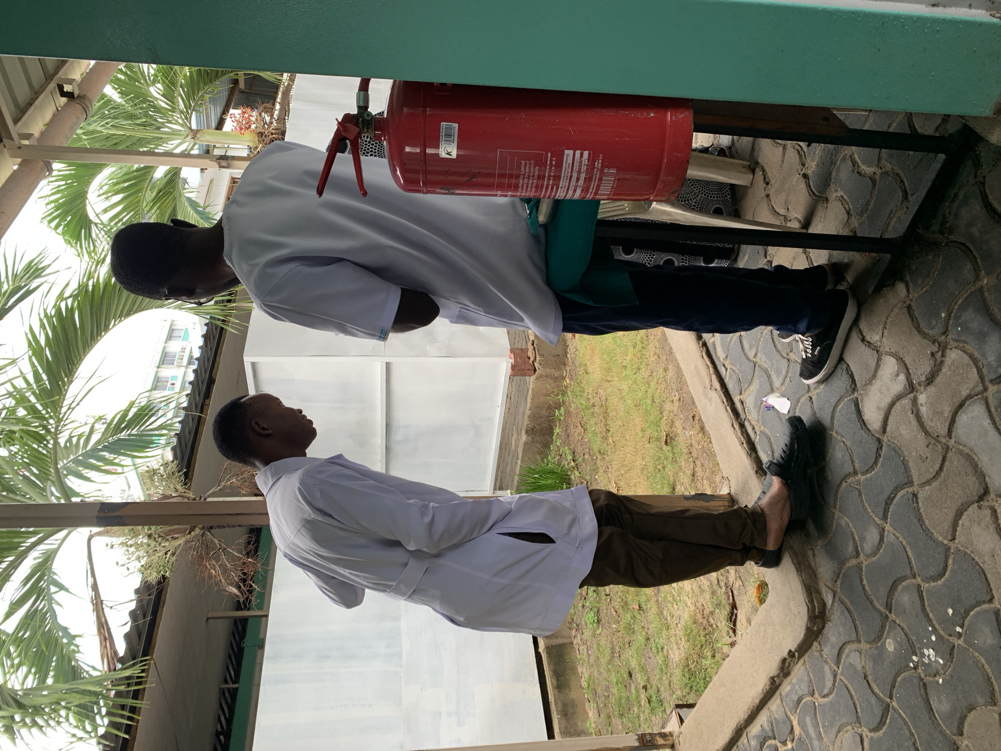 The image depicts two adult males interacting in an outdoor, partially sheltered area in an urban environment consistent with Dar es-Salaam, Tanzania. A precise location within the city is not discernible from the provided visual information, though a "Property of Zakea Medical" label on a fire extinguisher suggests proximity to a medical facility.
The ground in the foreground is paved with dark grey interlocking concrete pavers arranged in a wavy pattern, separated from a patch of green grass by a concrete curb. A light-colored structural pillar supports an overhead corrugated metal roof.
The individual on the left, a male with dark skin and short dark hair, wears a light lavender long-sleeved lab coat over dark brown trousers and black clogs worn with exposed heels. He is bent forward at the waist, looking down towards the pavement, with his hands in his coat pockets. A small, crumpled white object rests on the pavement near his left foot.
The individual on the right, also a male with dark skin, short dark hair, and wearing glasses, stands with his back mostly to the camera. He wears a white short-sleeved top, dark blue trousers, and black sneakers with white soles. A red cylindrical fire extinguisher, labeled "Property of Zakea Medical" and bearing the identifier "WM 0003132," stands prominently on a black metal frame to his right. A dark green fabric is draped over an unseen object situated between this individual and the fire extinguisher. A partial white chair or stool is visible behind the extinguisher.
The background features a plain white wall or partition. Above this, lush green palm fronds are visible, and in the distance, a multi-story building with light-colored walls and windows is partially obscured by vegetation. A teal-colored structural element frames the right side of the image. The scene is illuminated by natural daylight.