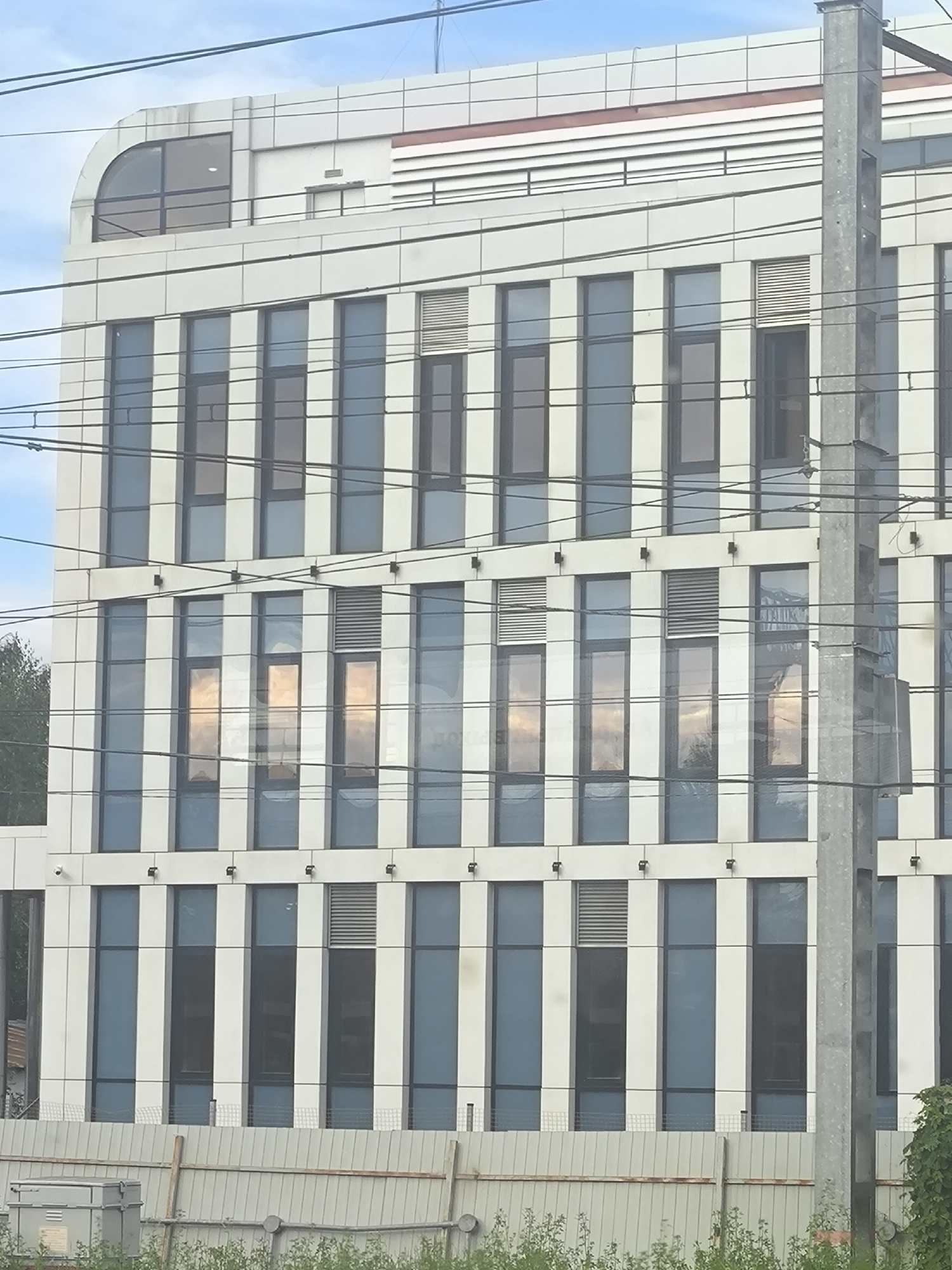 This media file is a photograph of a modern building with a facade of white panels and large, dark-framed windows. The building has multiple stories, and the windows are arranged in vertical sections. Some of the windows reflect the sky and clouds, creating a warm, golden hue on the facade. There are also horizontal louvers visible on some sections of the building, suggesting ventilation or architectural features.
In the foreground, there are utility wires stretching across the frame, obscuring parts of the building and implying an urban or semi-urban setting, possibly near a transportation route. A gray utility box is visible in the lower left corner. In the background, a few trees are partially visible on the left side, peeking out from behind the building. The sky is blue with scattered clouds, indicating daytime.
There is no discernible activity or event taking place. The scene appears to be a static view of the building and its surroundings. No people are visible. The overall mood is neutral, focusing on the architecture and the play of light on the reflective surfaces. No text is visible in the image.