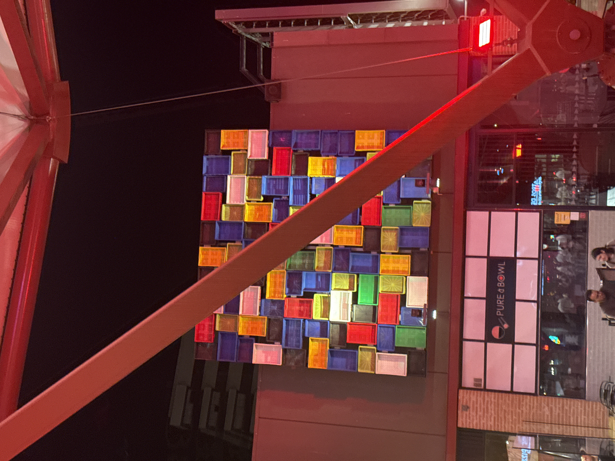 The image shows an outdoor scene at night, with a large, colorful art installation made of plastic crates mounted on a building facade. The crates are arranged in a grid pattern and display a variety of vibrant colors including red, blue, yellow, green, orange, and white. A diagonal red beam runs across the foreground, partially obscuring the art installation. Below the art installation is the storefront of a business named "PURE & BOWL". The storefront has large windows reflecting the activity inside, and several people can be seen inside and outside the establishment. The building facade is a mix of brick and modern materials. The overall impression is of a lively urban environment at night, with a focus on the artistic display and the commercial activity. The image is not taken in Alcorcón, Spain.