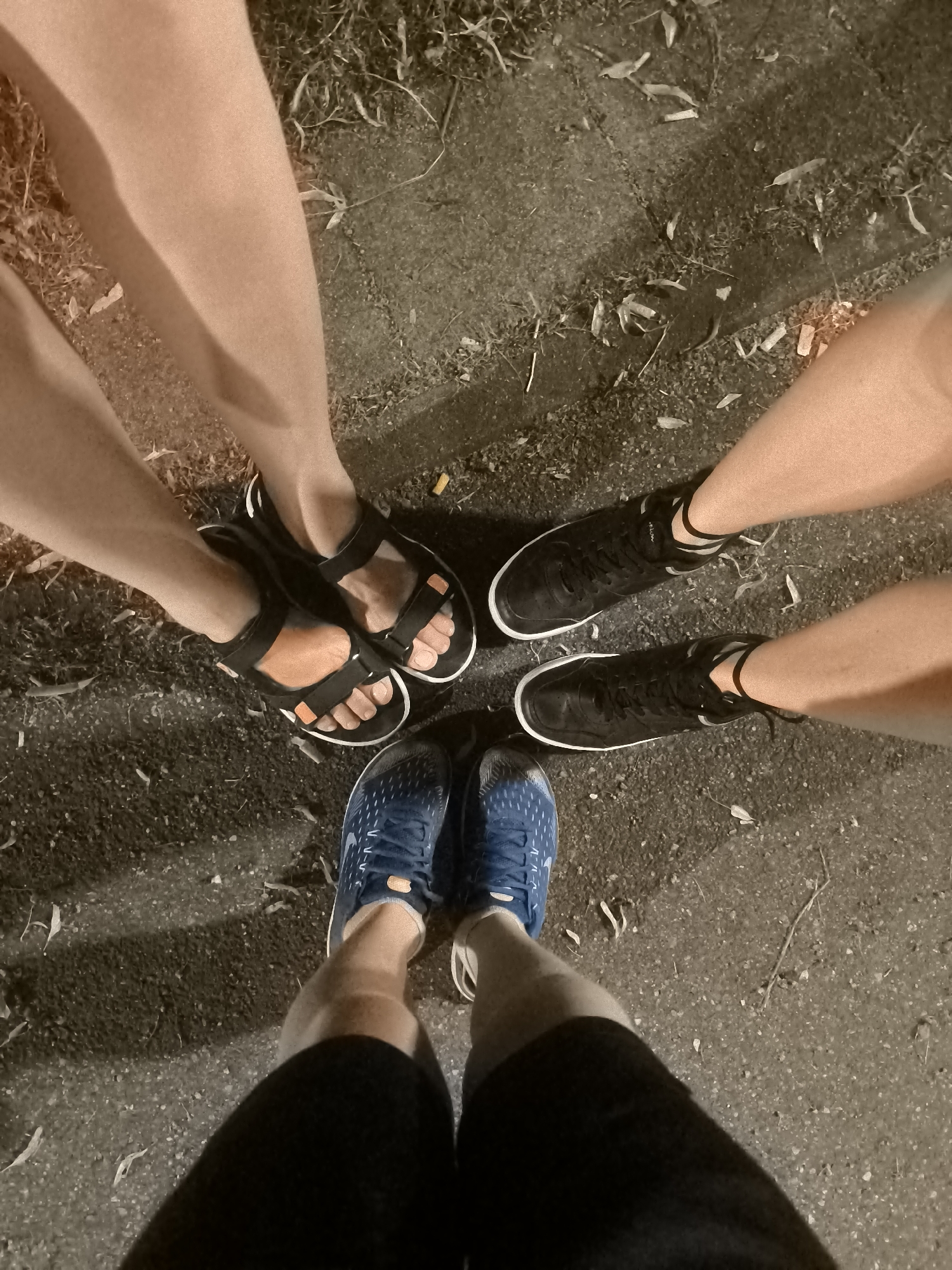 A group of four people stand in a circle facing outwards, their feet visible in the foreground. They are standing on a paved surface with scattered leaves and debris. The individuals are wearing a variety of footwear, including black sandals with orange accents, black athletic shoes with white soles and trim, and blue athletic shoes with white soles and laces. The lighting suggests it may be evening or night. The scene is likely outdoors in an urban or semi-urban environment, but the exact location is unknown.