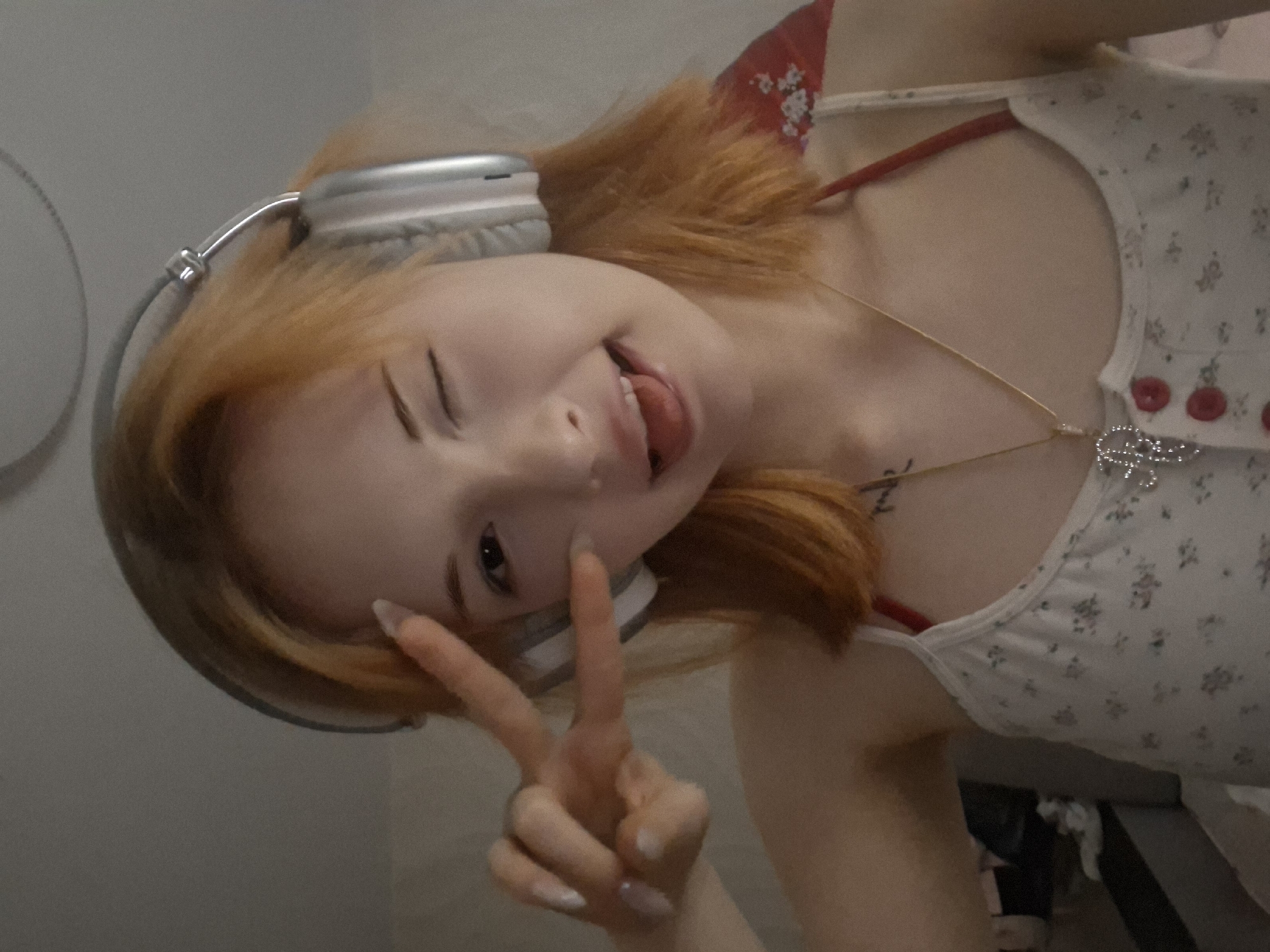 A young person with light skin and reddish-orange hair is shown in a selfie-style image. They are wearing white headphones and a light-colored tank top with a floral pattern and red straps. The person is making a peace sign with their hand, winking with one eye, and sticking out their tongue. They have a gold necklace with a pendant. The location is unknown and there is no evidence it is in Chaikovsky, Russia.