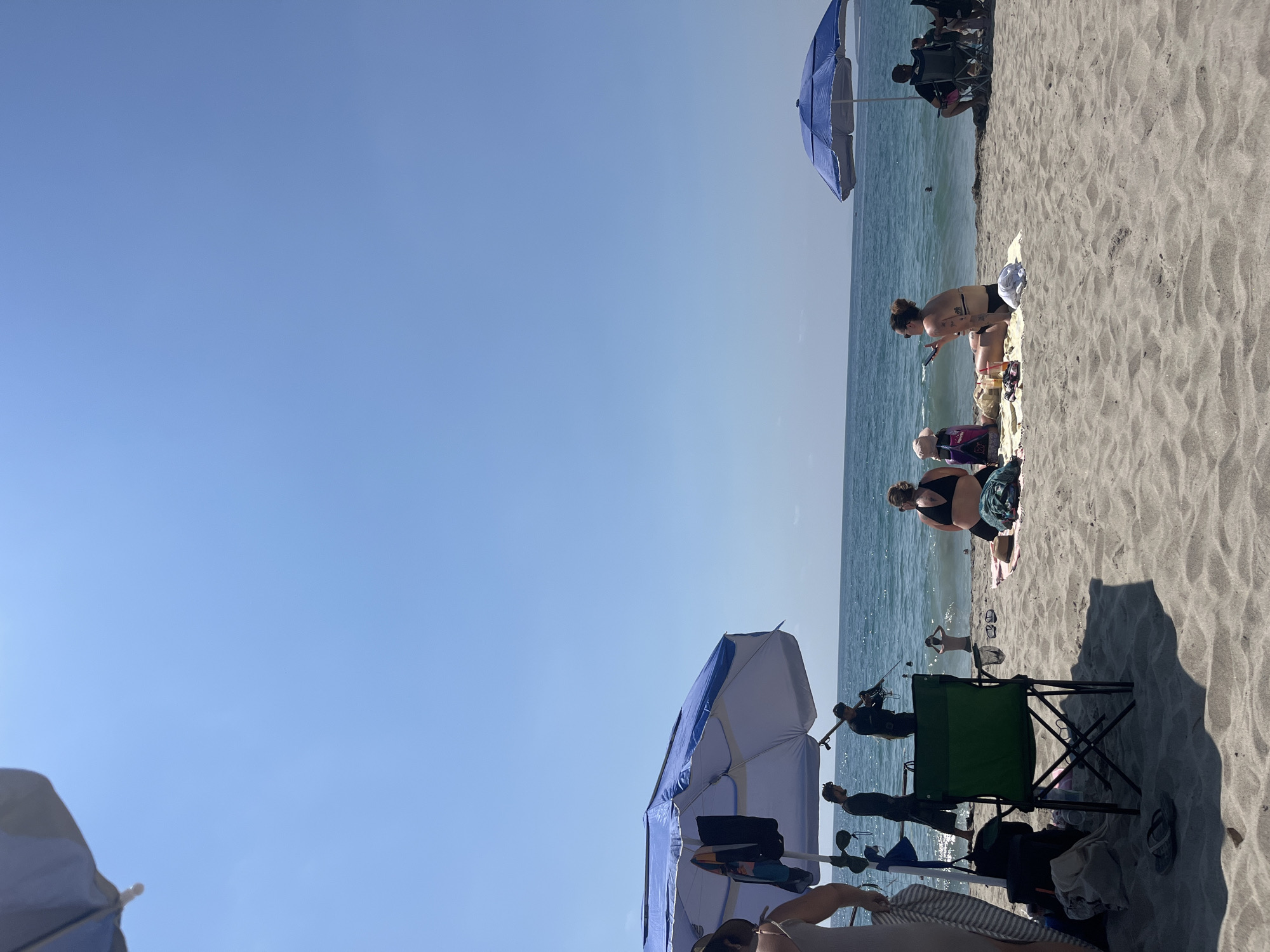 The image shows a beach scene in Brisas de Zicatela, Mexico. The beach is sandy, with a few people enjoying the sunshine. Some individuals are standing near the water's edge while others are sitting on towels or chairs. Several blue beach umbrellas provide shade, casting shadows on the sand. A green folding chair sits next to an occupied beach umbrella. The water is calm and the sky is clear.