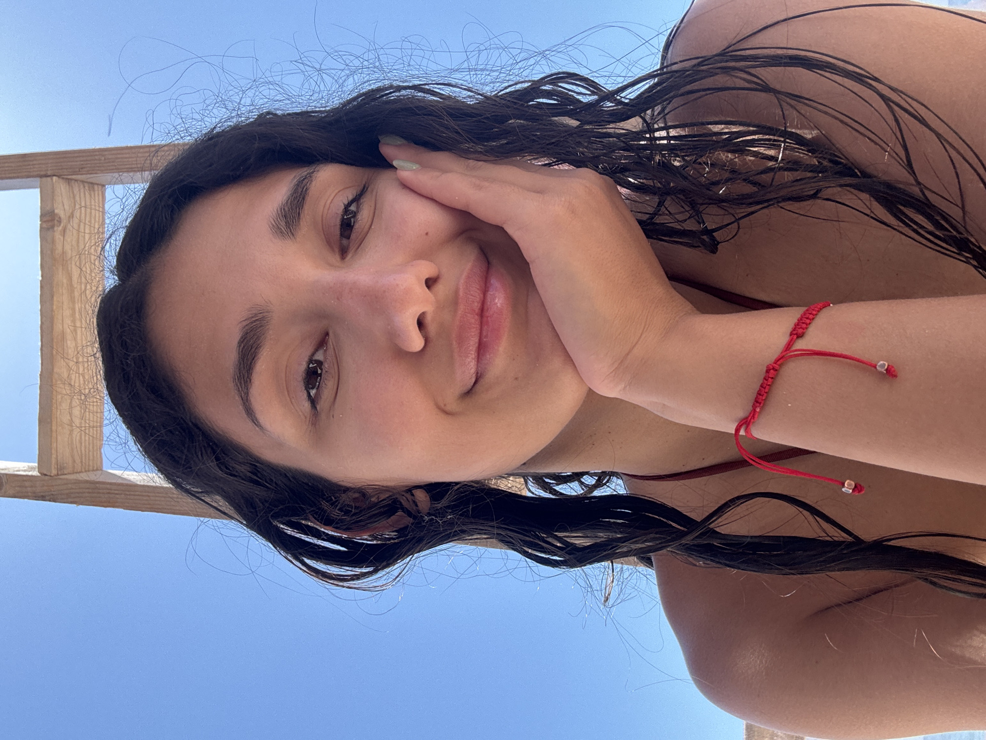 A close-up, upward-angled selfie features a young female subject reportedly in Vrba, Montenegro. The subject has a light to medium complexion and dark, wet, wavy hair. She is smiling, looking directly at the camera, and has her left hand resting against her left cheek. Her fingernails are painted a light green. A red string bracelet with two small silver beads is visible on her right wrist, and a red bikini strap is partially visible over her left shoulder. The background primarily consists of a clear blue sky, with a partial view of a light-colored wooden structure visible in the upper right quadrant. The scene is illuminated by bright daylight.