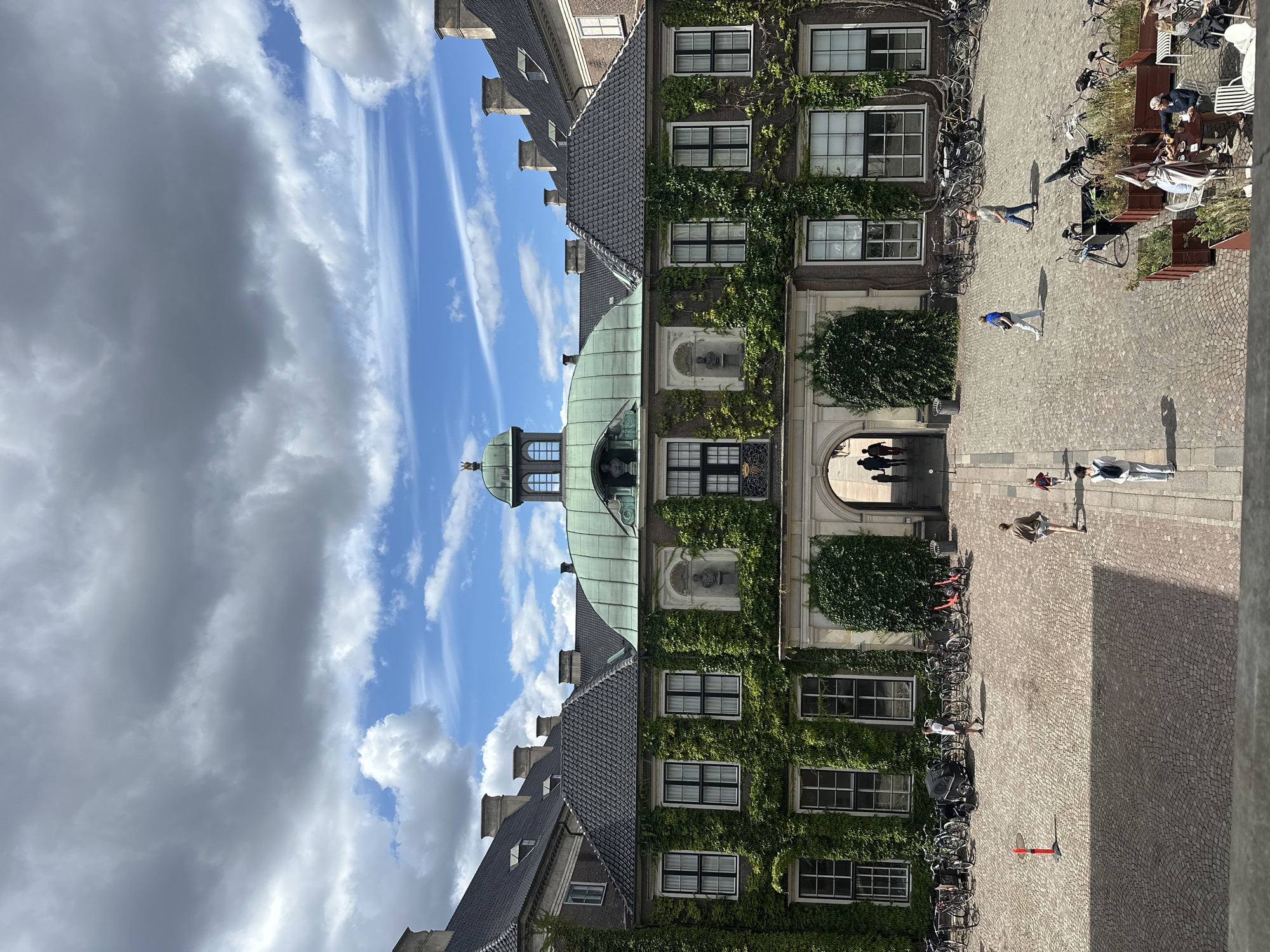 The image captures a courtyard in Copenhagen, Denmark, likely within Rosenborg Castle, evidenced by the architectural style, greenery, and courtyard layout typical of historical Danish buildings.
In the foreground, a cobblestone courtyard is partially covered by a shadow. Several people are walking through the courtyard. Towards the right, a group of people are seated at tables outside what appears to be a cafe.
In the mid-ground, a row of bicycles and a baby stroller are parked against a building. The building itself is a historical structure with a dark tiled roof and an ivy-covered facade. It features multiple windows with white frames. A large arched entrance is visible in the center of the building, leading into another area.
The background reveals more of the historical building, including a green domed roof with decorative elements and a small statue. The sky above is partly cloudy, with a mix of dark and light clouds against a blue sky. The overall scene depicts a sunny day in a historical European city.