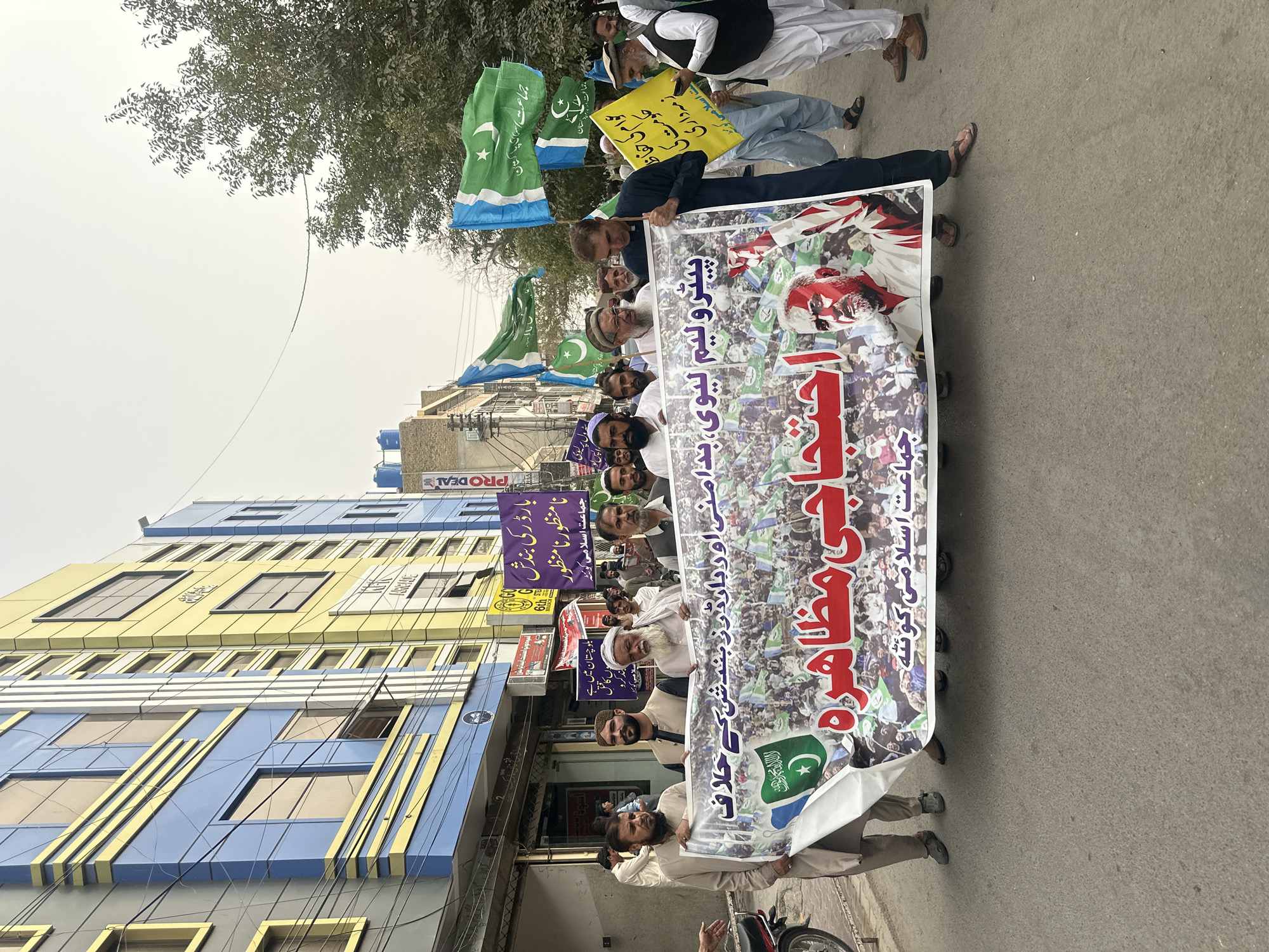 A group of people are marching in the city of Quetta City Tehsil, Pakistan, holding a large banner and several flags. The banner, with red and white text and an image of a man with red paint on his face, reads "احتجاجی مظاہرہ" (protest demonstration) and "پیٹرولیم، ہیوی، ودیگر بندش کے خلاف" (against the blockage of petroleum, heavy, and others). The flags are predominantly green and white, with a crescent moon and star symbol, characteristic of the Pakistani flag, and some feature slogans in Urdu. The people are dressed in traditional Pakistani attire, including shalwar kameez and kurtas. Some individuals are holding the banner from the top, while others are walking alongside. In the background, there are buildings with blue and yellow facades, as well as trees and an overcast sky. The street appears to be paved with asphalt. The overall scene suggests a political or social protest.