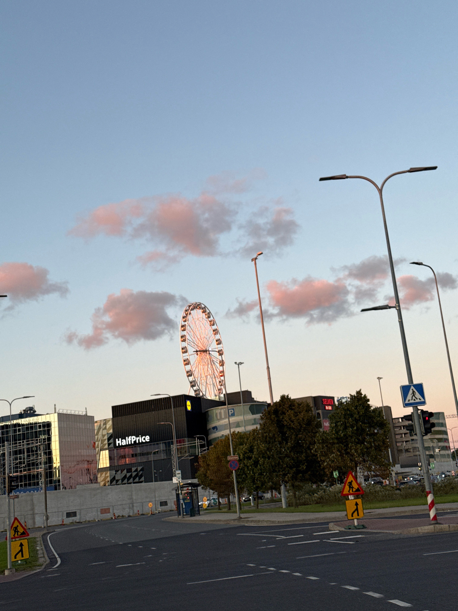 The image shows a street intersection in Tallinn, Estonia, with a large Ferris wheel in the background. The sky is a pale blue with scattered pink clouds, suggesting it is either early morning or late afternoon. Several streetlights are visible, and a pedestrian crossing sign is present. Buildings line the street, including a structure with a "HalfPrice" sign. There are traffic signs indicating road work and a detour. The road is asphalt with white lane markings. No people are clearly visible in the frame.