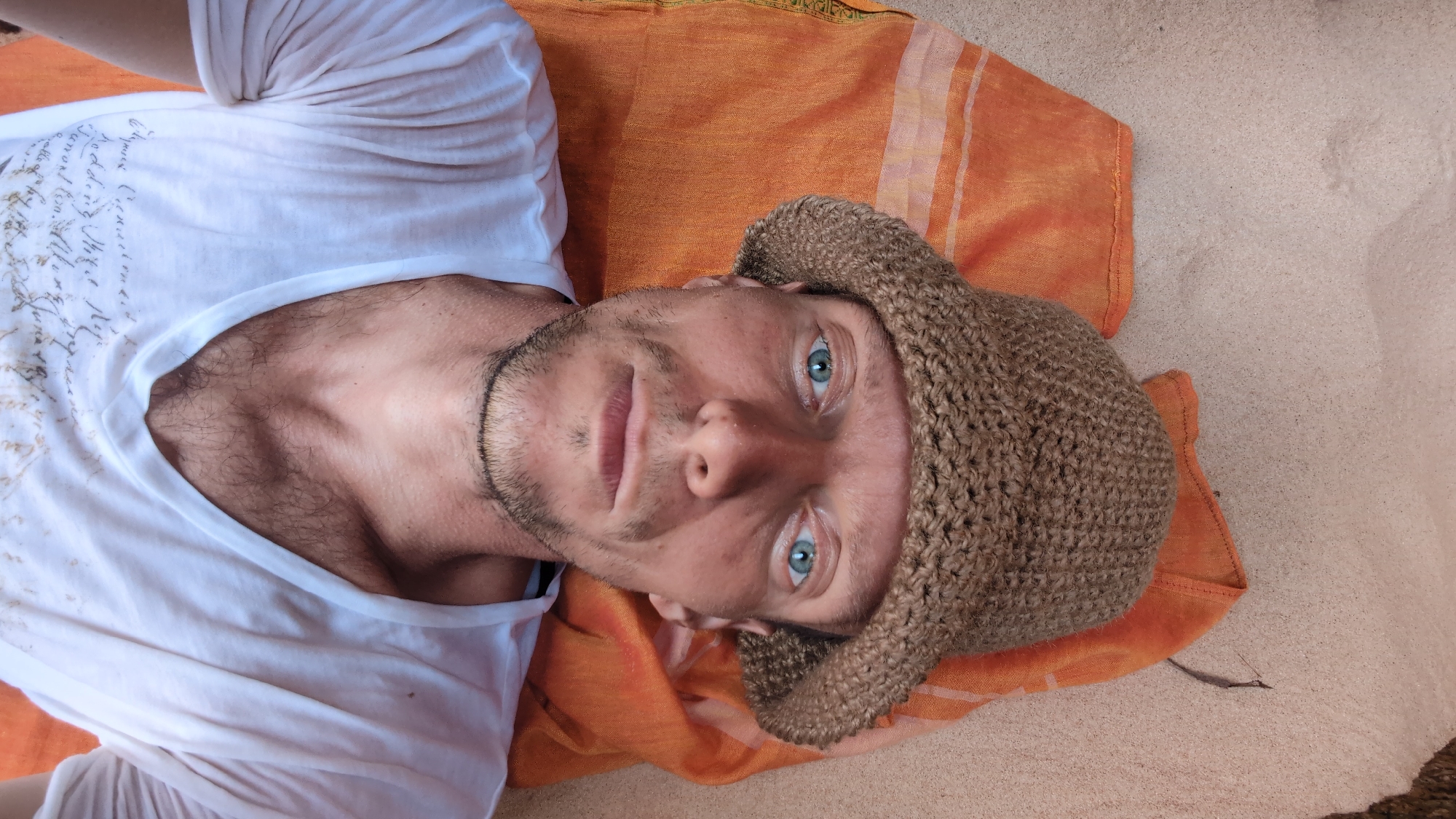 A light-skinned male with blue eyes, a short beard, and a brown knit cap lies on an orange patterned cloth. The cloth rests on sandy ground. He wears a white t-shirt with designs on the front. The scene is outdoors. The location is Phuoc Dinh, Vietnam.