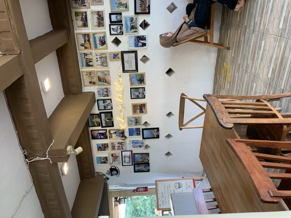 A wooden table and four chairs are arranged in a corner of a cafe located in Kecamatan Borobudur, Indonesia at -7.606 latitude and 110.18 longitude. The walls are white and adorned with framed photographs, including a picture of the iconic Borobudur Temple. A neon sign that reads "Kedai & Restoran" is mounted above the photographs. A woman in a brown hijab sits on a wooden chair with a tan cushion. Her legs are crossed and she is looking down at her phone. The wooden floors have a tiled pattern. A fire extinguisher is visible in the corner of the room, and a menu board is leaning against the wall. There is a large window in the background that looks out onto a verdant garden. The cafe is brightly lit and has a relaxed ambiance.