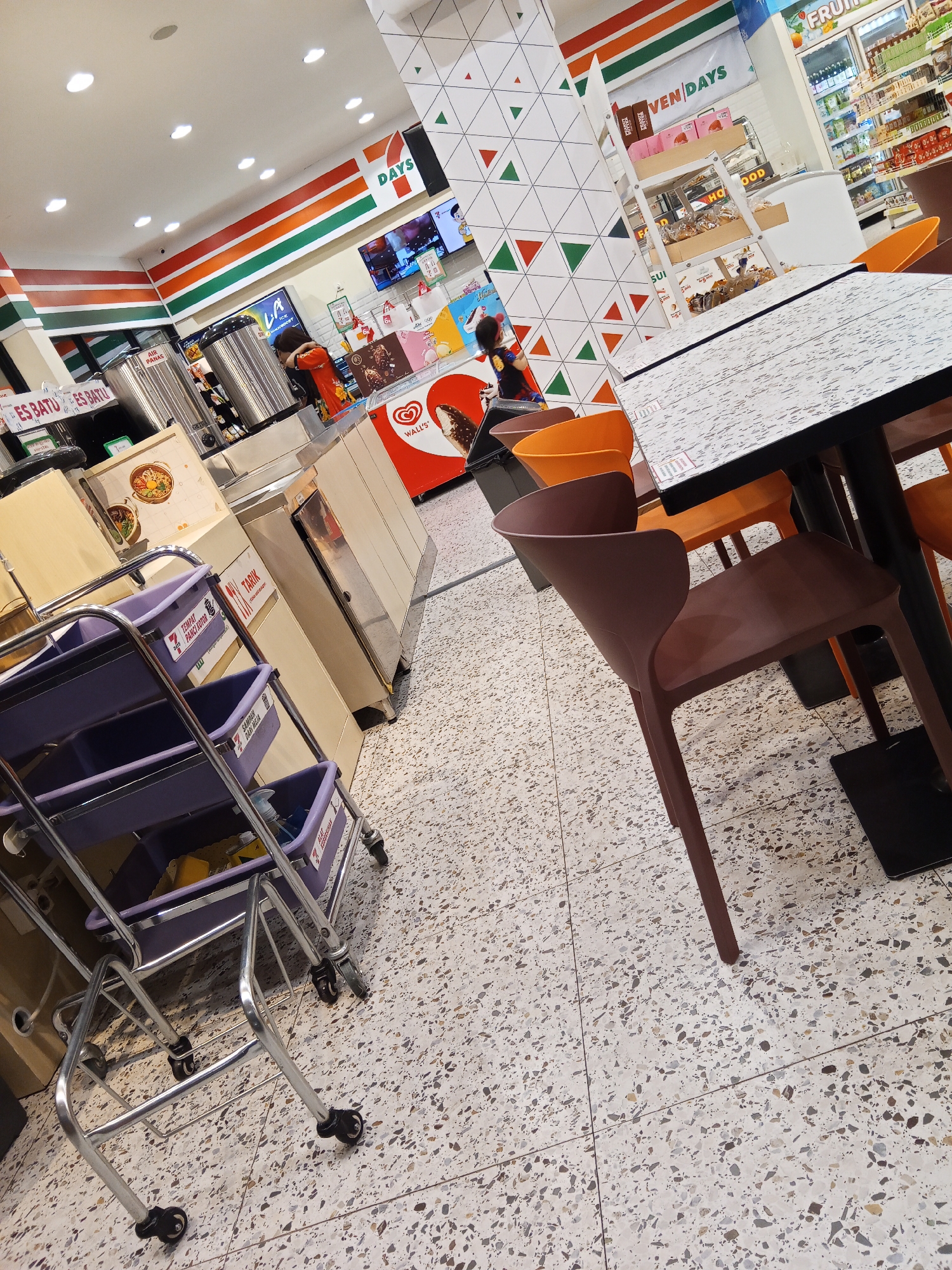 The image shows the interior of a 7-Eleven convenience store in Lubuklinggau, Indonesia. The store features the brand's signature orange, green, and red stripes on the walls. In the foreground, a metal cart with purple bins is positioned on the terrazzo-patterned floor. Beyond the cart, a dining area is visible with a table and several chairs, including a dark brown chair in the right foreground. In the background, shelves are stocked with various food and beverage items, and a large refrigerated display case is present. A sign displaying "7 DAYS" is mounted on the wall. The overall environment appears to be a typical retail space.