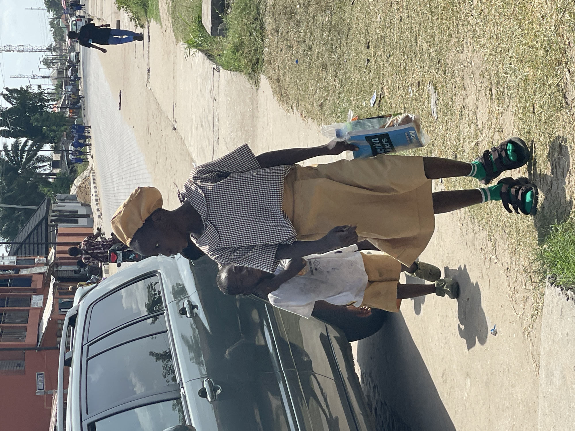 In Badagry, Nigeria, two figures, likely school children in uniform, are standing on a sidewalk next to a dark SUV. The older figure, wearing a checkered shirt, khaki skirt, hat, green socks, and sandals, holds a package of "Alton Colòn Filters". A younger child, in a white shirt and khaki shorts, stands next to the older one. The SUV's dark windows reflect the surroundings. Behind the SUV, people are walking on the road. In the background, a street scene unfolds, with buildings, cars, and power lines visible.