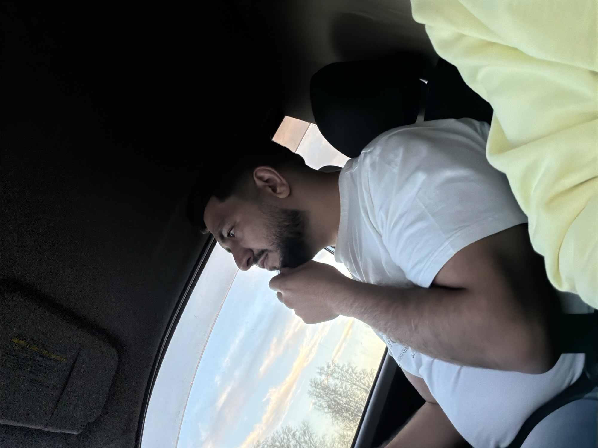 A medium shot from inside a vehicle facing a man in a white T-shirt who is looking out the window. He has dark hair and a beard, and he is touching his chin. Outside the window, the sky is visible, and there are trees in the distance. A light yellow cloth is draped over the passenger seat and part of the man’s shoulder. The sun visor is down on the driver's side, and the car interior is mostly dark. The location and city in Russia cannot be determined from this image.