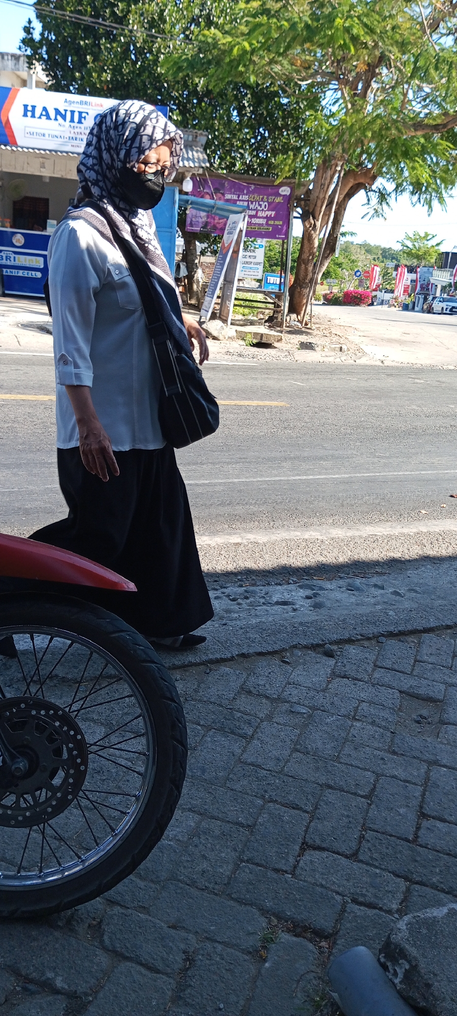 In Pakkanna, Indonesia, a person wearing a patterned hijab, glasses, and a face mask walks along a paved sidewalk next to a street. The individual is dressed in a light-colored, three-quarter sleeve shirt and a long black skirt. A dark bag with a strap is slung over their shoulder. To the left, a red motorcycle is partially visible, with its front wheel and spokes in the foreground. Behind the person, a building with signage for "AgenBRIlink HANIF" is visible. Further down the street, a tall tree with lush green foliage dominates the background, casting shadows on the ground. Other structures and red and white flags are present in the distance, suggesting a commercial or residential area. The sky is clear and bright.