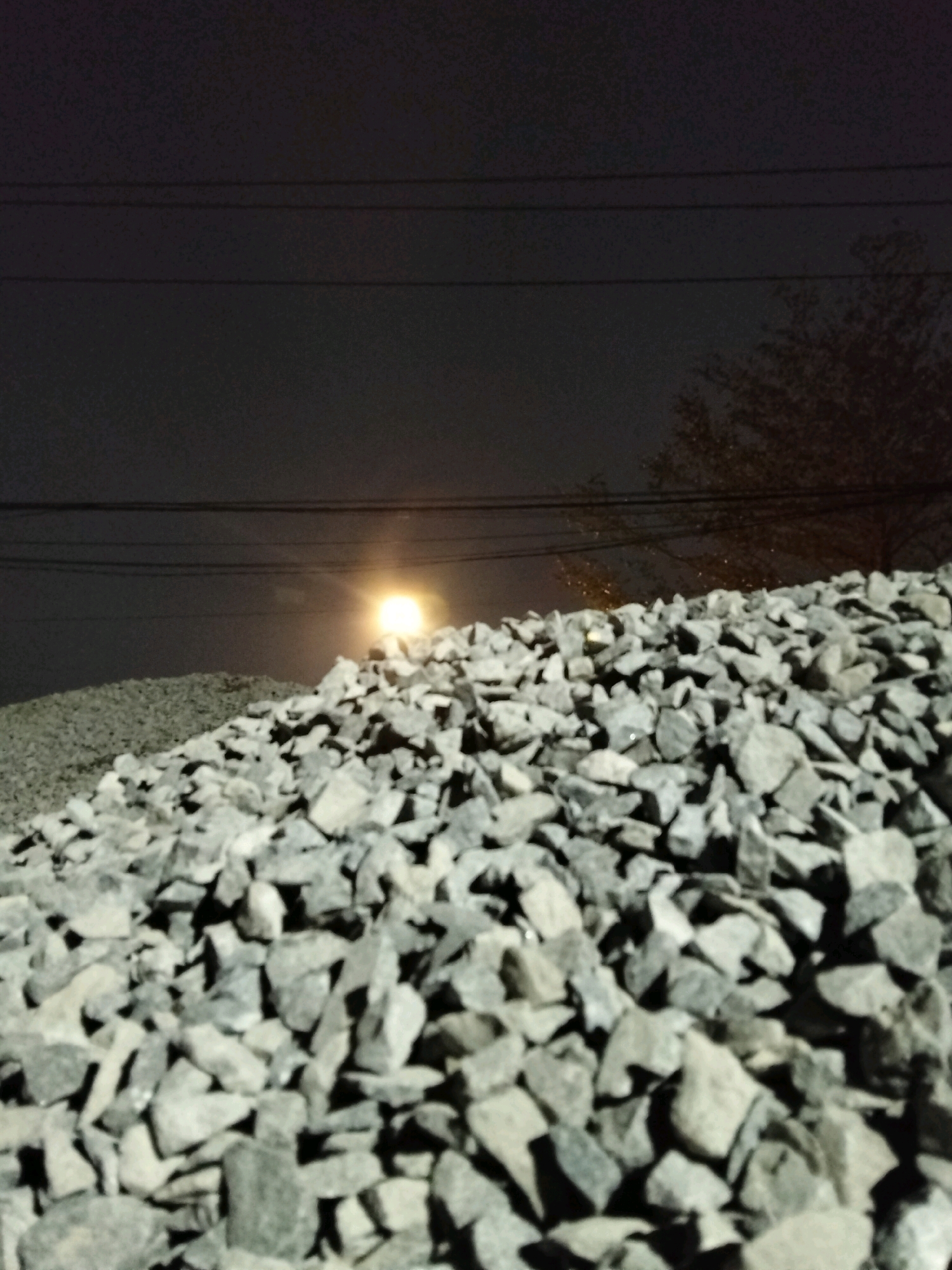 The image displays a nighttime scene with a pile of gray gravel in the foreground. In the background, power lines are visible against a dark sky, and a faint, glowing light source, possibly the moon or a distant streetlight, is positioned above the gravel. A silhouetted tree is faintly visible on the right side of the frame. The overall environment suggests an outdoor location, possibly a construction site or a roadside area. There are no people or specific landmarks visible that would allow for pinpointing the location within Pontianak, Indonesia.