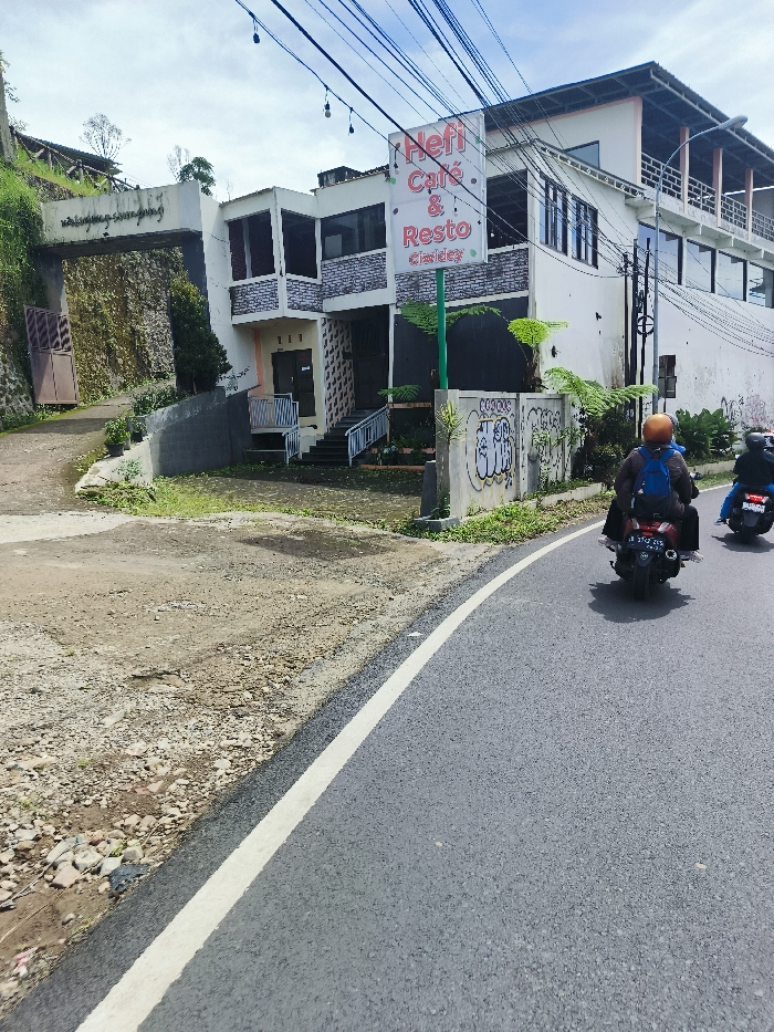 The image shows a scene in Rancabali, Indonesia, located at coordinates -7.12 latitude and 107.432 longitude. A two-story building with a sign reading "Hefi Cafe & Resto" is located on the right side of the image. The building has a white exterior with large windows and a balcony. A white painted curb runs along the edge of a paved road. A person wearing a blue shirt and helmet is driving a black motorbike towards the camera. Another person wearing a blue shirt and helmet is driving a black motorbike away from the camera. The motorcycle is traveling on a paved road. The road is lined with a white painted curb. There is a concrete ramp leading up to the building. Vegetation is located on the right side of the building. Vegetation is located on the left side of the image. The sky is clear and blue with white clouds. The pavement is in good condition, but the area to the left is covered in gravel.