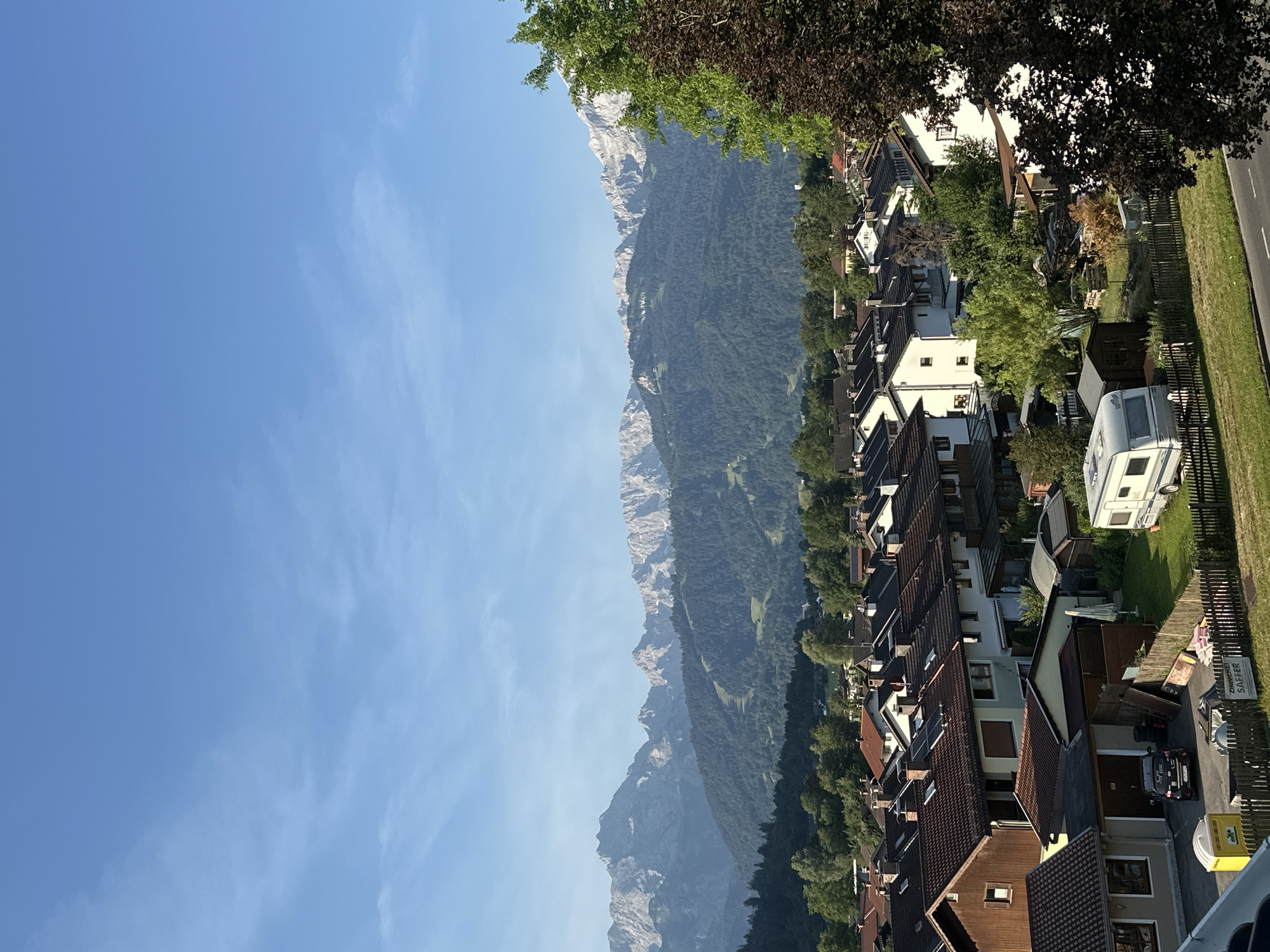 The image shows a residential area in Garmisch-Partenkirchen, Germany, with a backdrop of majestic mountains. The foreground is filled with a cluster of houses, characterized by their brown and white facades and dark tiled roofs. Some of the houses have solar panels installed, indicating modern energy solutions. A few yards are visible, with some featuring wooden fences, garden sheds, and neatly mowed lawns. There's a white caravan parked on a grassy area. A dark colored car is parked in a driveway next to a garage. A portable toilet is also visible. In the middle ground, more houses and trees create a sense of a developed neighborhood. The background is dominated by lush green hills covered with dense forests, leading up to a dramatic range of snow-capped mountains under a clear blue sky. The lighting suggests it is daytime, likely morning or late afternoon, given the soft illumination and the shadows cast. No people are visible in the frame.