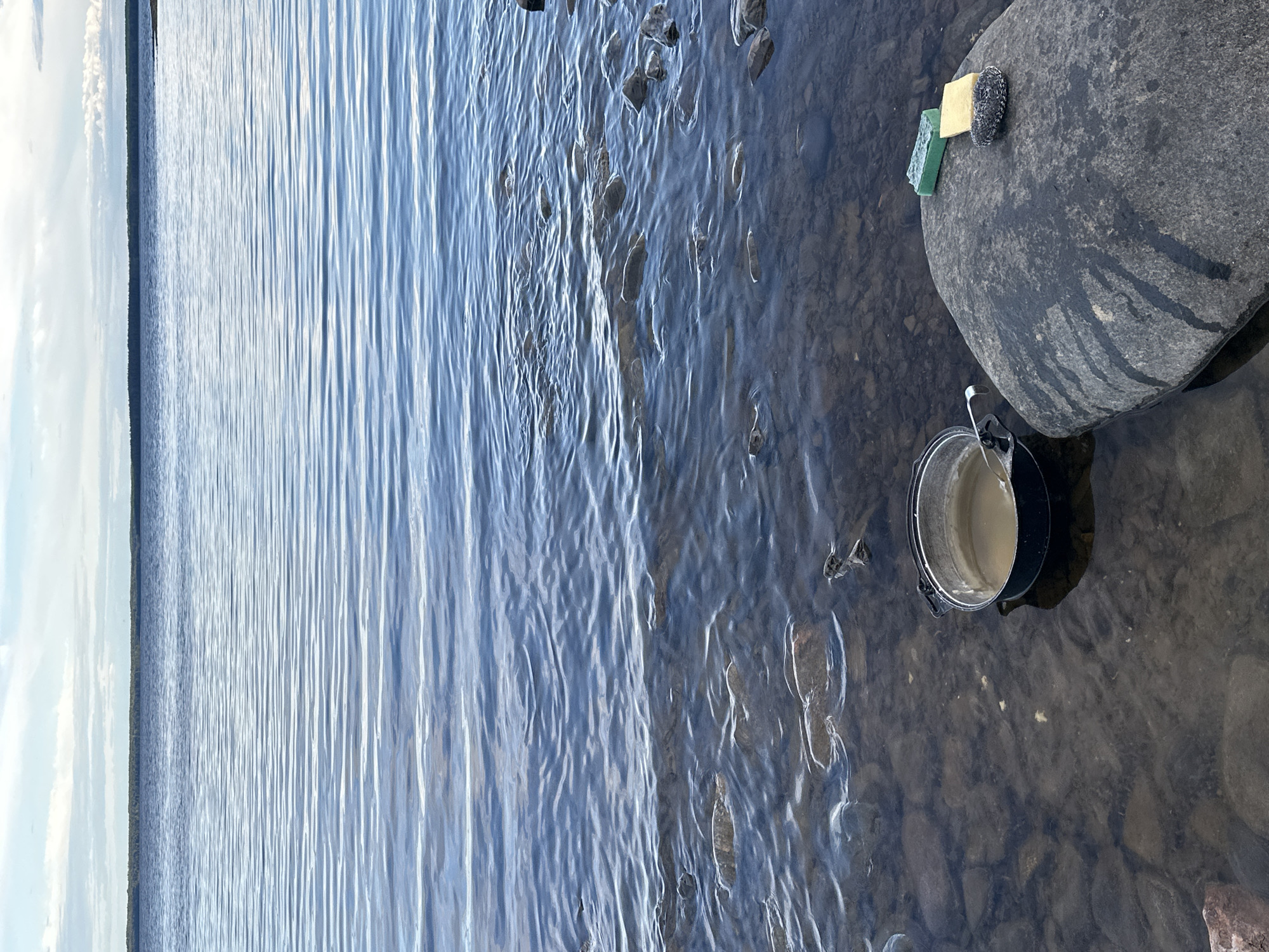This is a vertical, medium-shot, outdoor, eye-level photograph taken on a sunny day. The scene depicts the shore of a body of water, likely a lake or calm sea, with the water occupying the left two-thirds of the frame and the shore occupying the right third. The water is a deep blue with ripples reflecting the bright sky, creating a shimmering effect. Along the shoreline, submerged rocks are visible beneath the clear, shallow water.
On the right side of the frame, a large, dark, wet rock is partially submerged. Resting on top of this rock are three cleaning sponges: a green rectangular one, a yellow square one, and a round gray and black scrub pad. Further to the right and slightly below the rock, a small, dark pot with a handle is sitting in the shallow water. The pot contains a light-colored liquid, possibly water mixed with mud or sand. The surrounding area is a rocky, pebbly shore. The overall impression is one of a serene natural setting, possibly being used for cleaning or preparation for an outdoor activity. The lighting suggests it is daytime, likely late morning or early afternoon, given the bright, diffused sunlight. No people are visible in the frame. There is no visible text in the image.