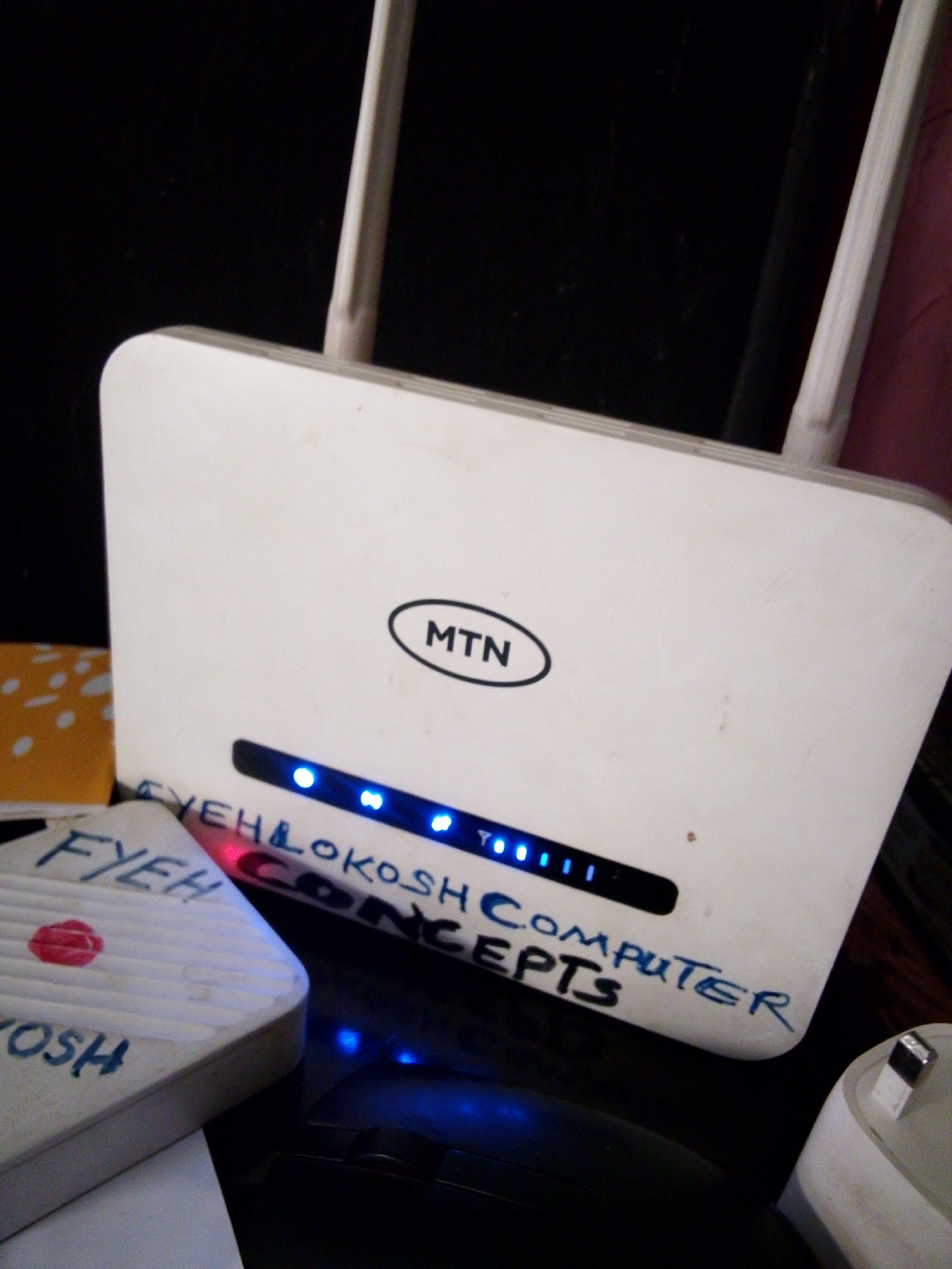 The image shows a white MTN broadband router with its antennas extended. The router has a prominent "MTN" logo in an oval shape. Below the logo, a strip of blue LED lights indicates network activity. There is handwritten text in blue marker on the front of the router. The visible text reads "FYEH LOKOSH COMPUTER CONCEPTS". To the left of the router, a smaller white object, possibly a router accessory or a small device, is visible with similar blue handwritten text "FYEH" and "OSH" along with a red circular mark resembling a lipstick print. The scene is dimly lit, suggesting an indoor setting, likely a home or office. The router appears to be connected and active, indicated by the lit LEDs. The handwritten text and the lipstick-like mark add a personal touch to the otherwise standard electronic device. The background is dark, making the illuminated LEDs and the white router stand out. The presence of the MTN logo suggests a telecommunications context. The overall impression is one of a functional electronic device with personalized markings, likely in a domestic or small business environment in Nigeria.