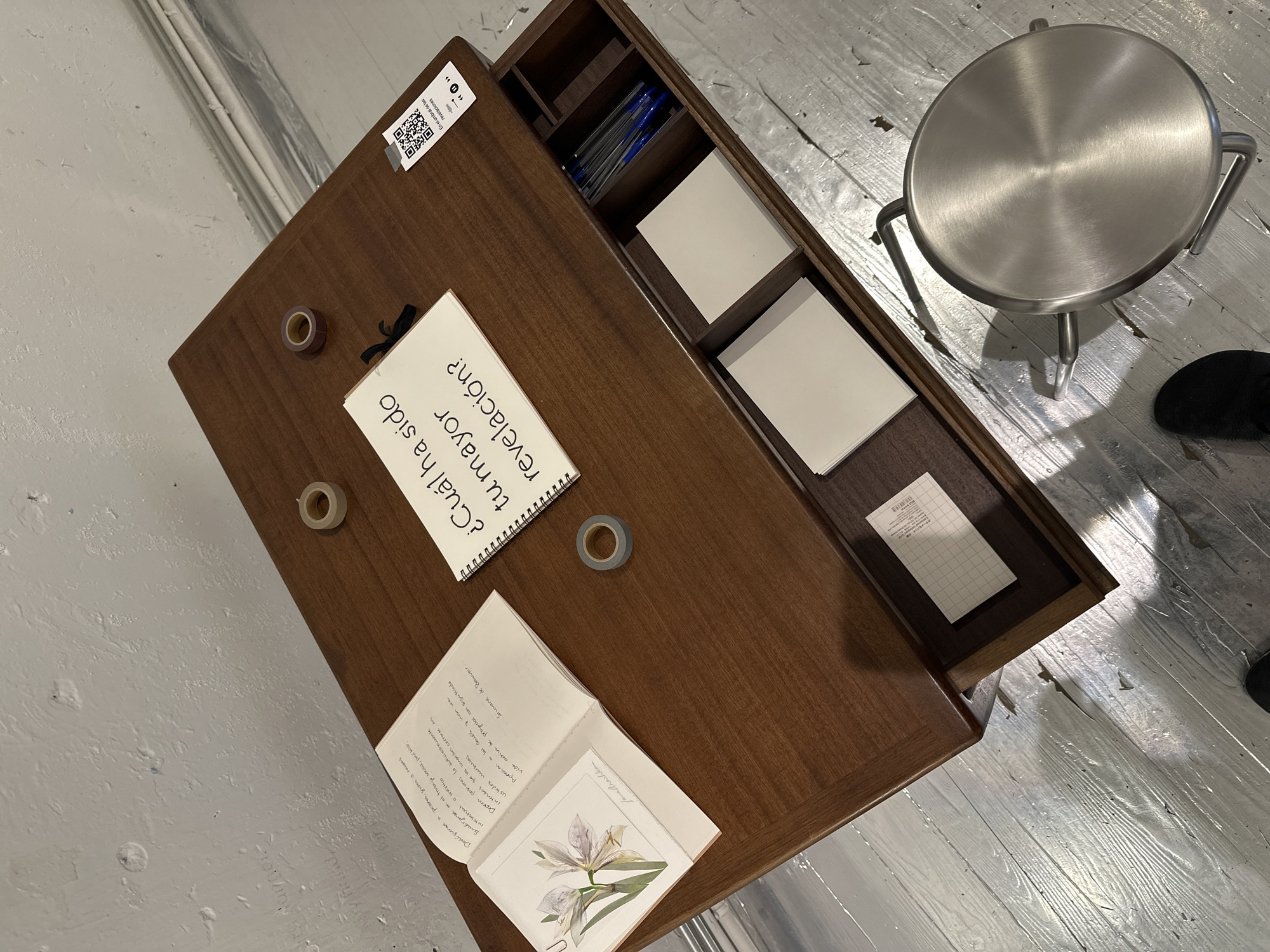 The scene is an art installation. A wooden desk with an open drawer contains supplies like pens, blank cards, and grid paper. A notebook on the desk poses the question, "¿Cuál ha sido tu mayor revelación?" (What has been your biggest revelation?). Another notebook is open with handwritten text in Spanish and a printed image of a white iris. The desk also has rolls of tape. A silver metal stool is positioned next to the desk, and parts of a person wearing black shoes are visible near the stool. The flooring is painted white with visible wood grain. A sticker on the desk states "En el umbral de las revelaciones" (On the threshold of revelations), indicating the theme of the installation. There is no evidence in the image that the scene is located in Madrid, Spain.