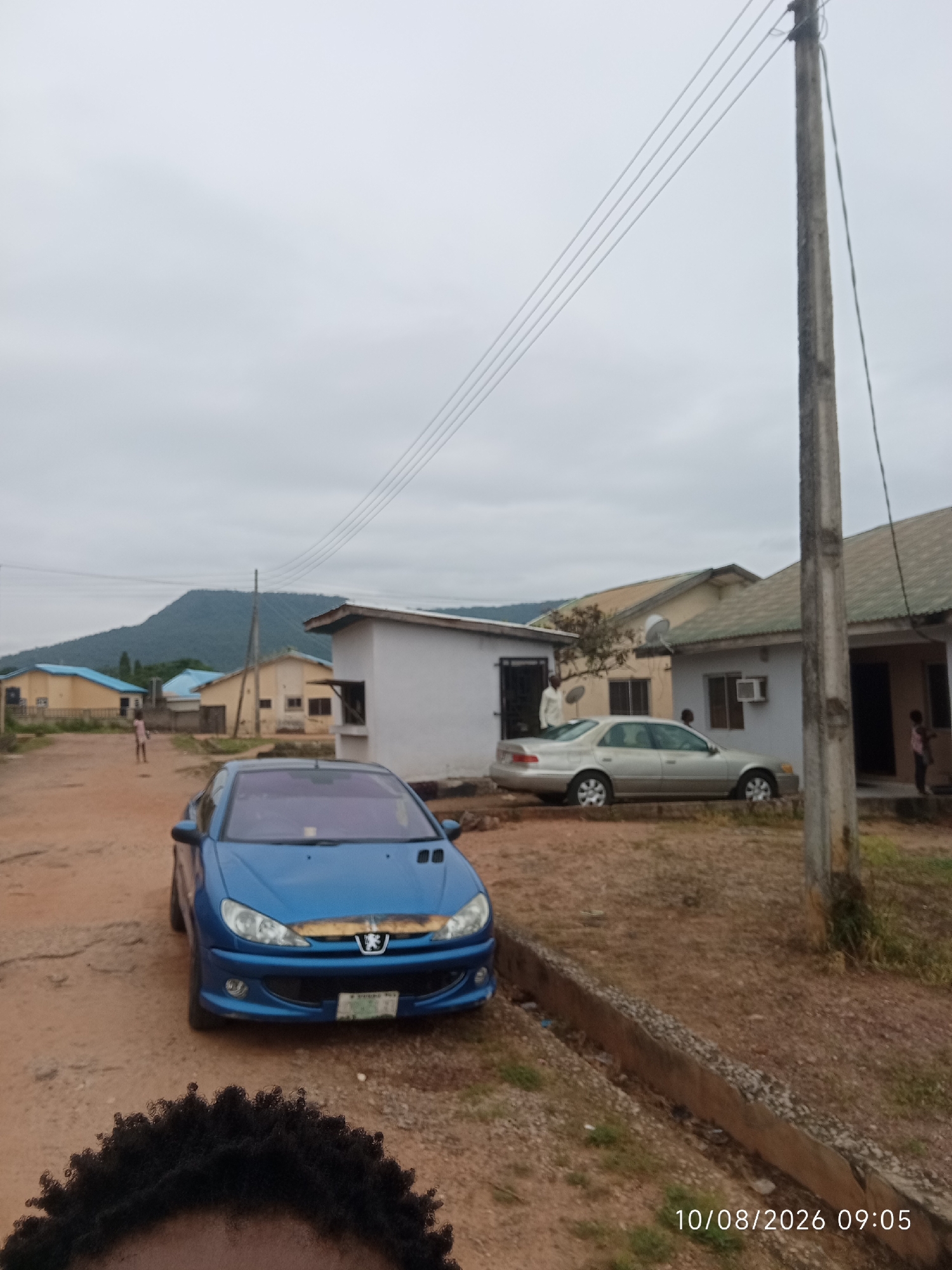 In Adankolo, Nigeria, on a dirt road under an overcast sky, a blue Peugeot 206 is parked. The car faces left, its headlights illuminated. Behind it, a silver sedan is parked in front of a light-colored building with a green roof. Further back, several other buildings with yellow and blue roofs are visible. A person is seen walking in the distance. A utility pole stands to the right of the frame, with power lines stretching across the sky. The timestamp "10/08/2026 09:05" is visible in the lower right corner. A concrete drainage channel runs along the right side of the road. A portion of a person's dark, curly hair is in the foreground at the bottom of the image.