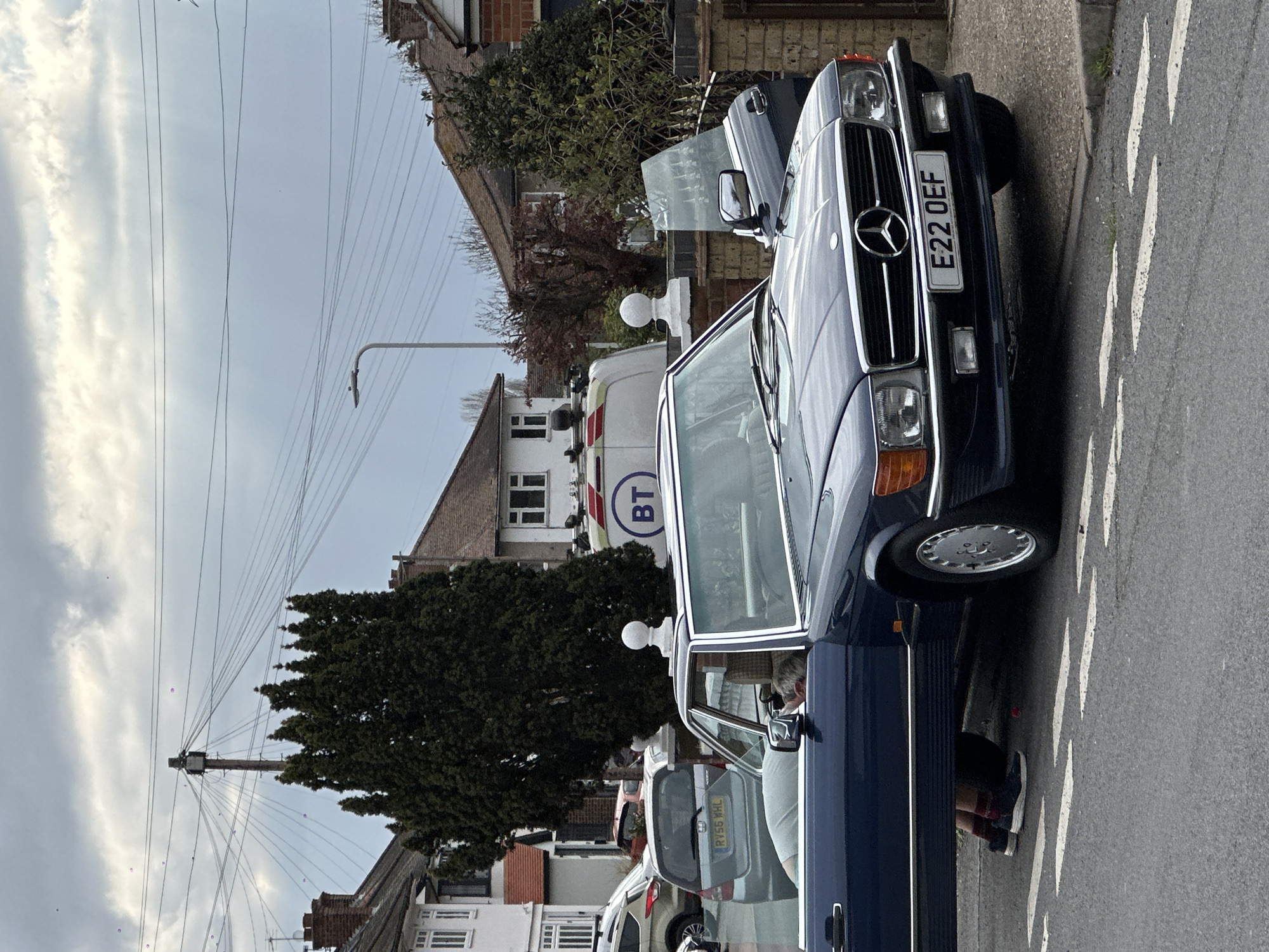 The image depicts a residential street scene in the United Kingdom, likely within the greater London area, under an overcast sky. A dark blue Mercedes-Benz car is parked at the curb. The car's license plate reads "E22 OEF." The driver's side door is open, and a person's lower body can be seen near the open door, wearing dark trousers, red and black socks, and white sneakers. Other parked vehicles are visible down the street, including a white BT (British Telecom) van and a gray car with a license plate beginning with "RY56." Houses line the street on the left side. Electrical cables stretch across the sky above the street. A lamppost is also visible. The road markings are white chevrons.