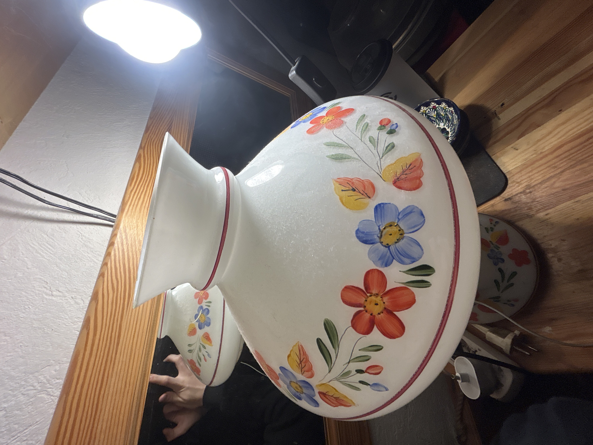 The image shows a close-up of a white, floral-painted lamp hanging from a ceiling. The lamp's shade features colorful flowers and leaves painted on its surface, and it has a decorative rim. The lamp is partially reflected in a mirror, revealing a person's hand and arm touching the mirror frame. The surrounding environment includes a wooden frame, possibly part of the mirror or a door, and a wooden surface or shelf in the background, upon which are several plates and a small ornate bowl. A light fixture is visible on the ceiling. The location appears to be indoors.