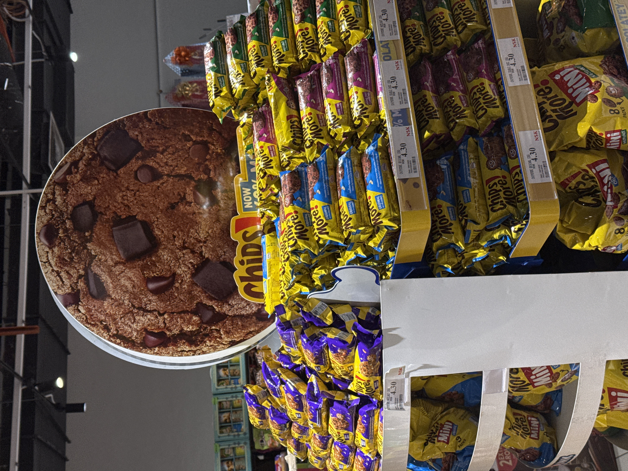 This media file depicts a retail setting, likely a supermarket aisle in Kuala Lumpur, Malaysia, judging by the presence of product labels in English and Malay, and prices indicated in RM (Malaysian Ringgit). The focal point is a display of snack products, primarily chocolate chip cookies and biscuit bars.
The products are neatly arranged on shelves and in hanging displays. Many of the packages feature the brand name "More!" and variations like "Chips!" and "Mini." Text visible on the packaging and shelf labels includes prices such as "4.30 RM" and product descriptions like "ORIGINAL," "HAZELNUT," and "DOUBLE DOC." Some labels also include codes and brand identifiers like "NSK."
In the foreground, a large circular advertisement for a chocolate chip cookie is prominent, showcasing a close-up of a cookie with visible chocolate chunks. The overall lighting suggests an indoor environment, possibly illuminated by store lighting, with no clear indication of time of day or weather. The scene is characterized by an abundance of commercially displayed goods, typical of a grocery or convenience store environment. There are no people visible in the frame.