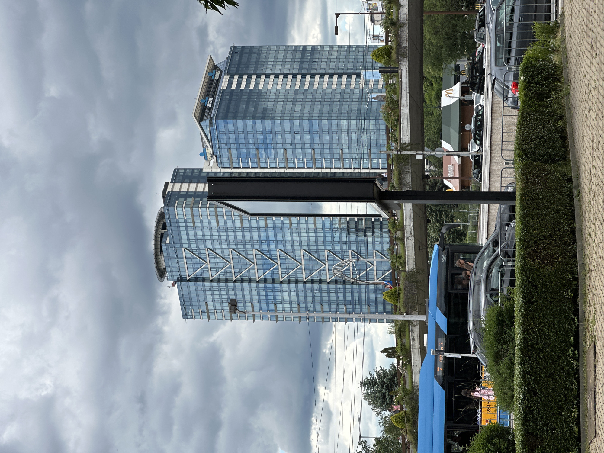 The image depicts a cloudy day in Sofia, Bulgaria, with a focus on two modern, glass-facaded skyscrapers. The larger skyscraper on the left features a prominent V-shaped geometric design on its facade and a helipad on its roof. The smaller skyscraper to its right displays corporate logos on its upper section. Below the skyscrapers, an elevated green area with landscaping and benches is visible. A blue bus with route number "7" is in motion on the left side of the frame, carrying passengers. In the foreground, a dark green hedge runs along the bottom of the image, with a paved area and metal railing beyond it. Several cars are visible on a road beneath the elevated greenery, and a McDonald's restaurant with its distinctive golden arches is situated further in the background. The overall scene suggests an urban environment with significant architectural structures and ongoing city life.