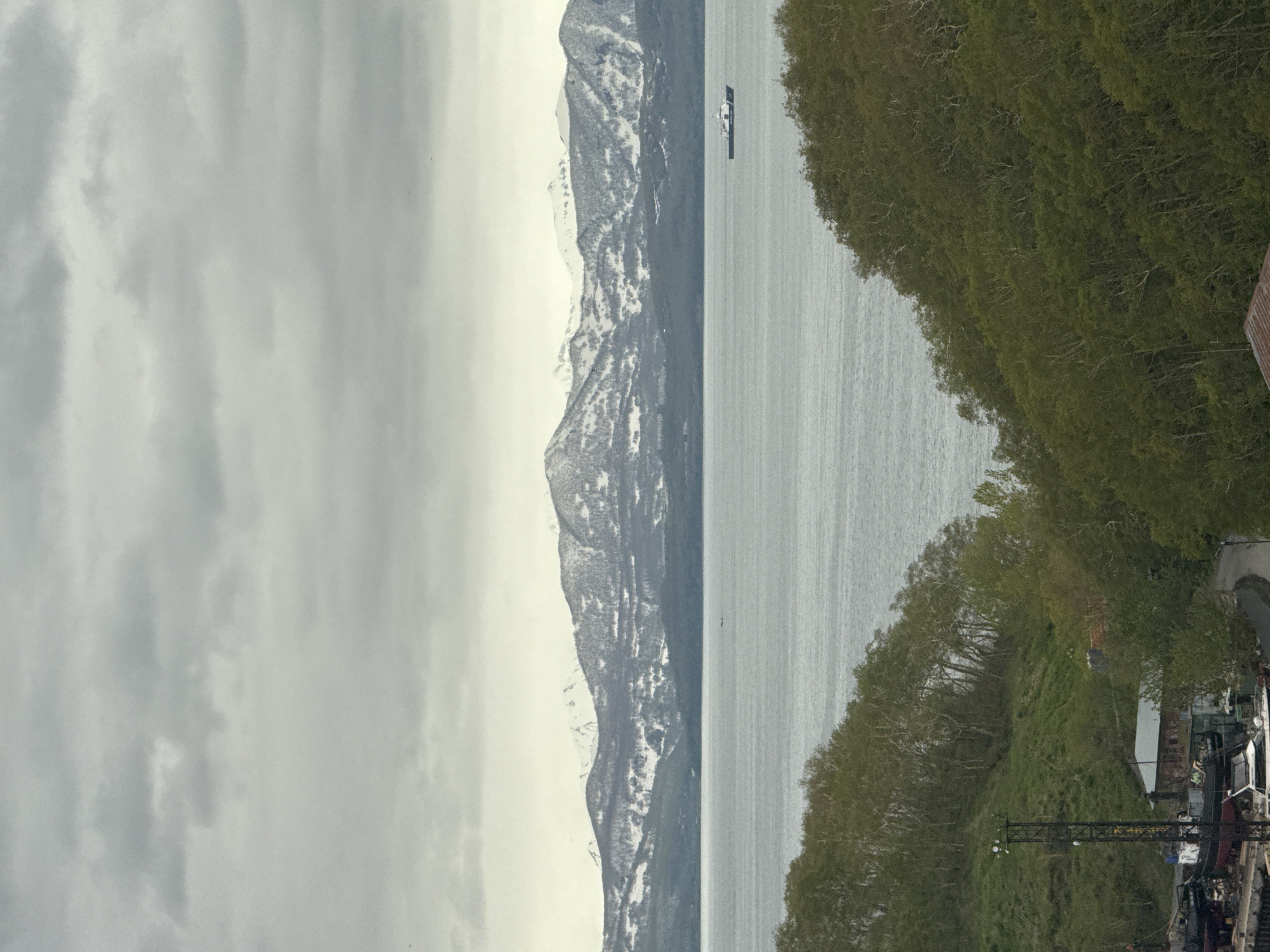 The image depicts a wide view of a body of water with a boat on it, framed by hills covered in trees. In the background, a snow-capped mountain range stretches across the horizon under a cloudy sky. In the foreground, a small industrial area is visible on the left, with boats on land and some small buildings. The overall scene suggests a coastal or harbor area in a region with significant elevation and a temperate climate.