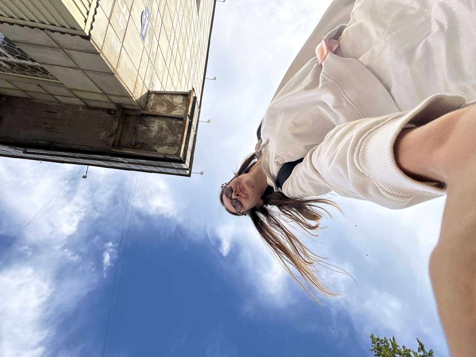 In Chaikovsky, Russia, under a bright blue sky with scattered clouds, a young woman is captured from a low-angle perspective, possibly taking a selfie. She is wearing a light-colored hoodie and sunglasses, with her long brown hair flowing. The background features the side of a building with a tiled facade and a distinct overhang, indicating an urban setting. The perspective is such that the building appears to loom above the subject, and the sky fills a significant portion of the frame. The image captures a candid moment, with the woman looking upwards and slightly to the side, her arm extended to hold the camera.