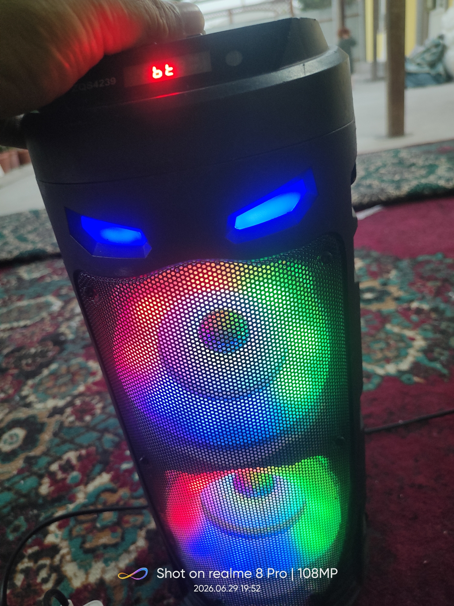 The image shows a close-up of a black portable speaker with blue LED lights resembling eyes and a colorful light show on its mesh speaker grills. A hand is visible in the upper left corner, partially obscuring a red "bt" indicator light and some serial numbers. The speaker is placed on a patterned rug, and the background suggests an indoor or semi-outdoor setting with blurry figures and structures visible. A watermark in the lower right corner indicates the photo was taken with a "realme 8 Pro | 108MP" on "2026.06.29 19:52". The presence of the specific watermark and date format strongly suggests this image is not from Bokhtar, Tajikistan, or the current time.