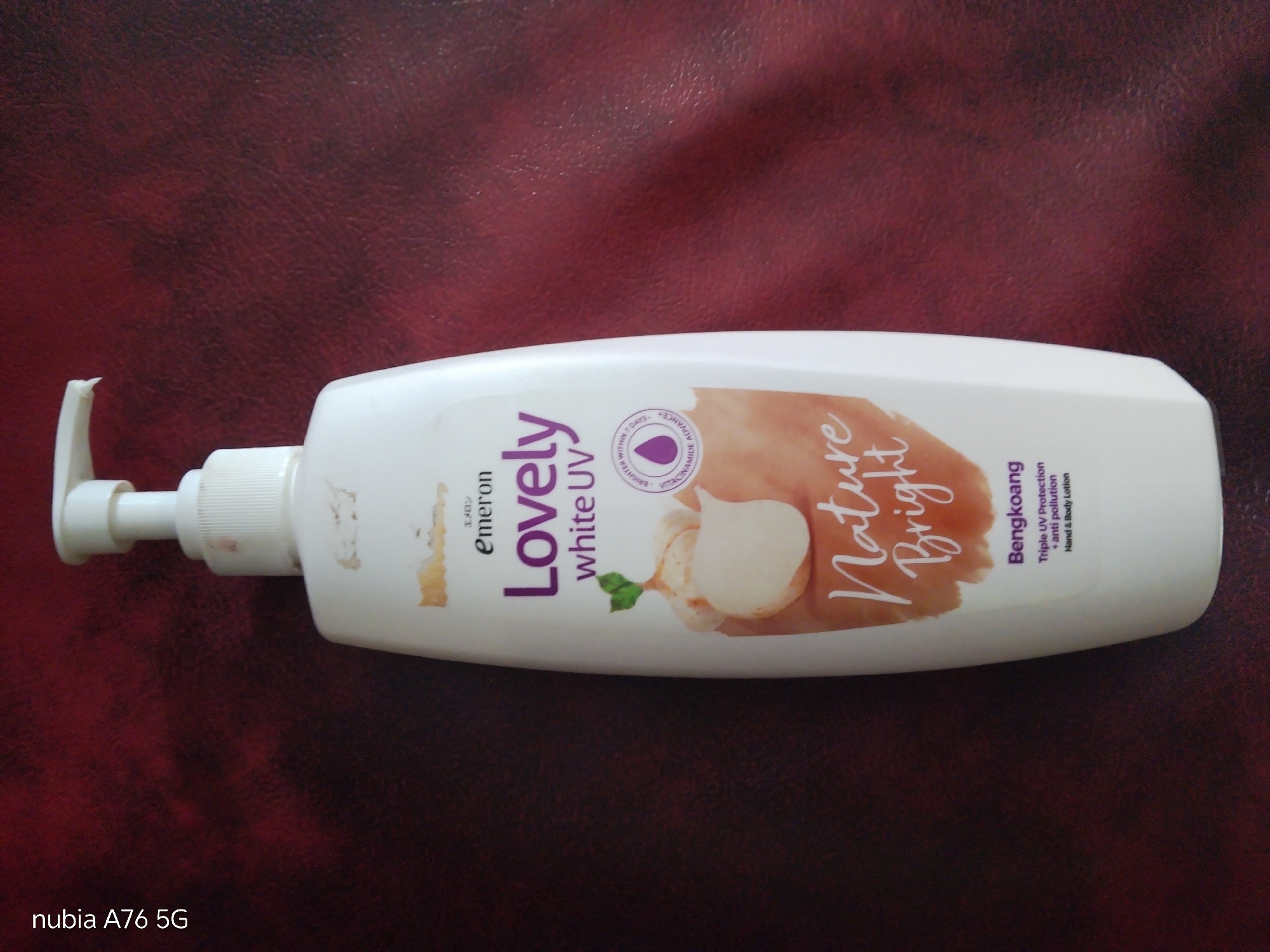 The image shows a white lotion bottle with a pump dispenser lying on a dark red, textured surface, likely a table or fabric. The bottle is positioned horizontally. The label on the bottle is predominantly white with purple and orange text and graphics. Prominently displayed are the words "Lovely" in large purple letters and "Emeron" in smaller purple letters. There is also text that reads "white UV" in smaller purple letters. Graphics on the bottle include an illustration of what appears to be ginger or garlic, along with a small green leaf. Further text on the label reads "Nature Bright" in orange script, and in smaller black text, "Bengkoang," "Triple UV Protection + anti pollution," and "Hand & Body Lotion." There is also a circular graphic with text indicating "BRIGHTER WITHIN 7 DAYS." In the bottom left corner of the image, the text "nubia A76 5G" is visible, indicating the device used to capture the photo. The overall lighting suggests an indoor setting. There are no people present in the image, and no specific location in Cakung, Indonesia, can be identified from the visual information provided.