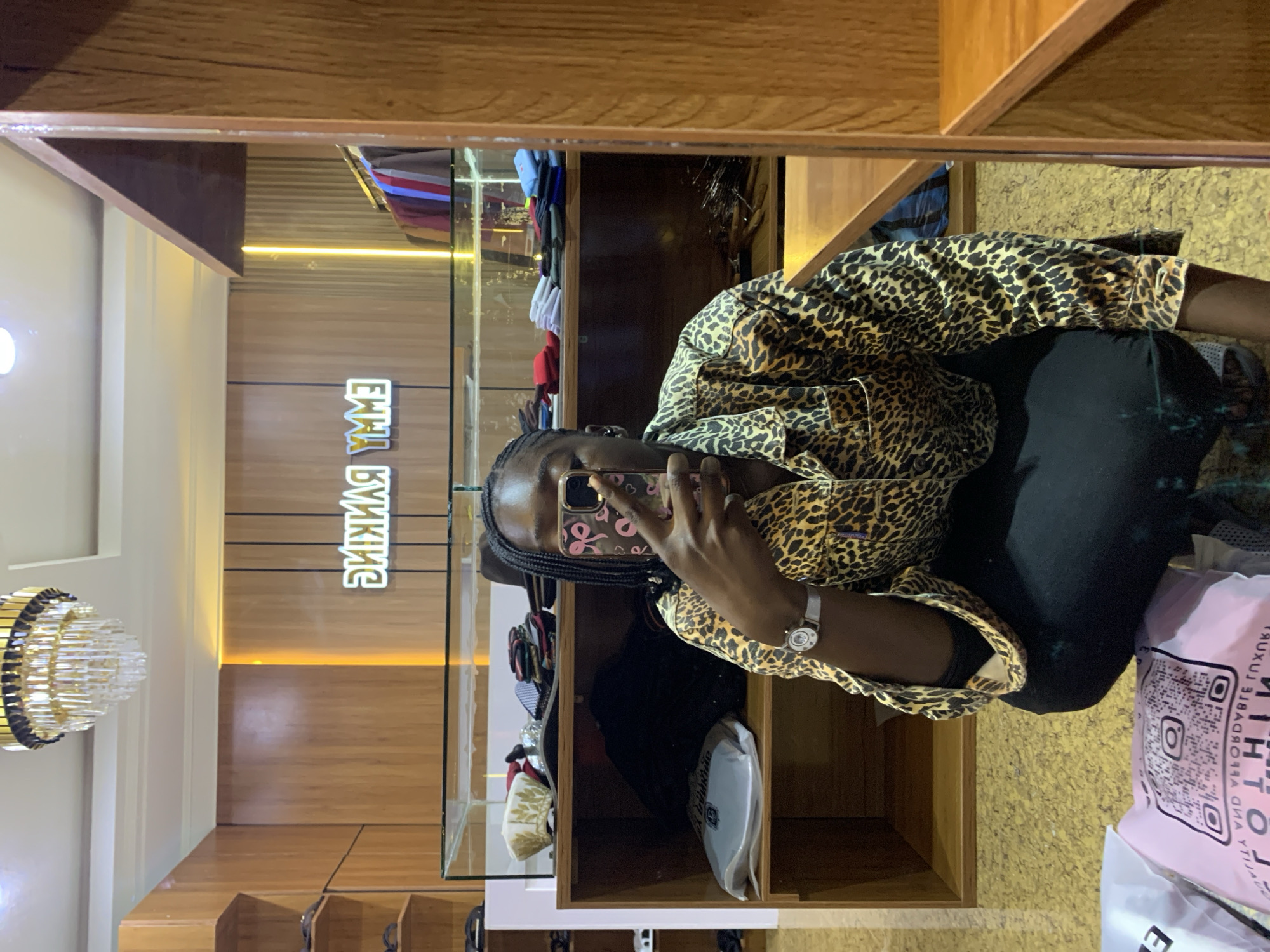 A young Black woman is taking a mirror selfie in a clothing store. She is wearing a leopard-print collared shirt and black pants. A watch is visible on her left wrist. She is holding a smartphone with a pink bow-patterned case up to take the photo. The store has wooden shelves displaying clothing and accessories. A neon sign that reads "EMMY RANKING" is visible on the back wall. The floor is a reflective yellow material. Shopping bags are on the floor in the foreground. The store appears to be located in Gura Top, Nigeria.