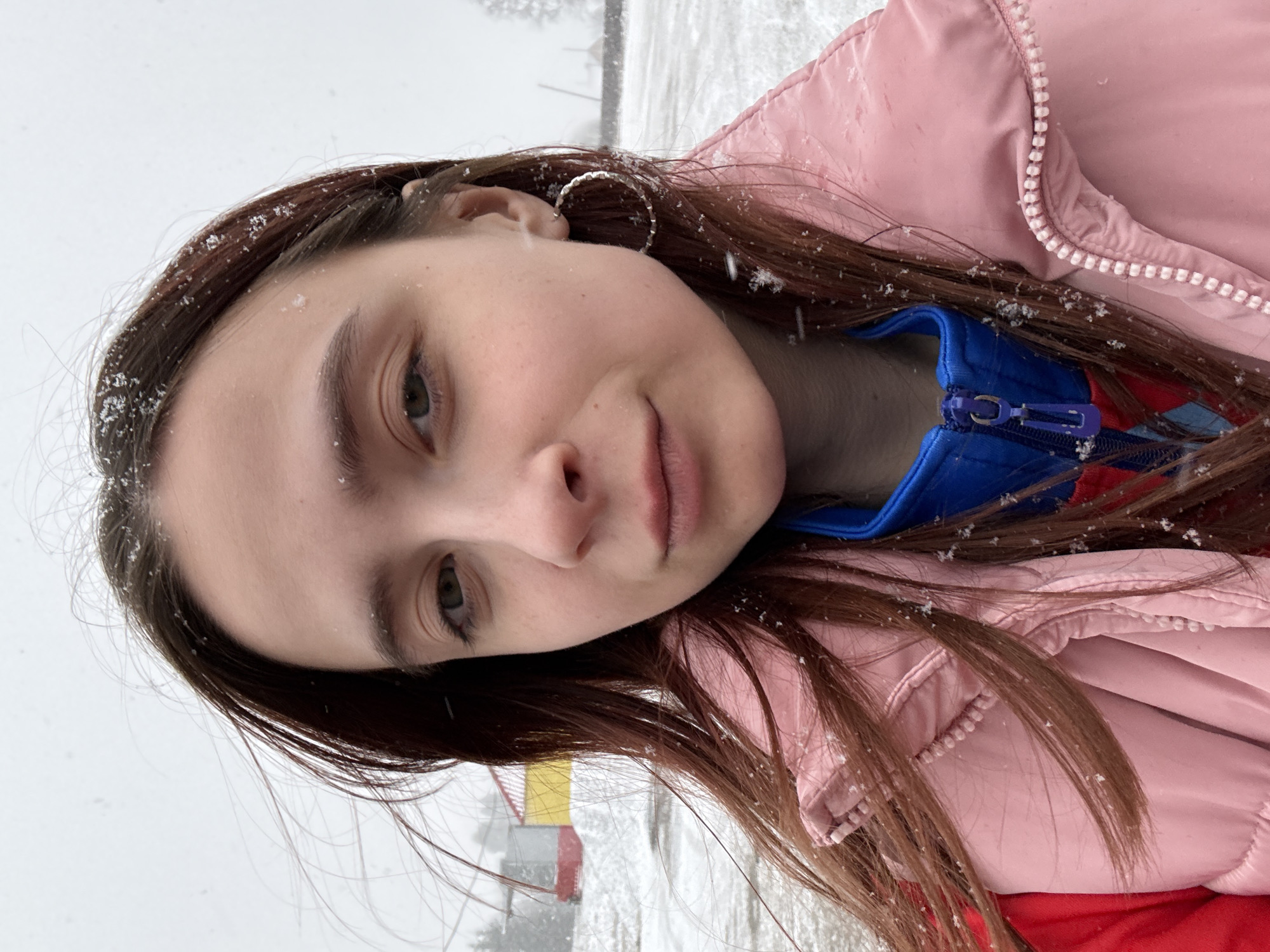 A young woman is pictured at an outdoor location near Gory, Russia. She is fair-skinned with brown hair, currently falling as snow, and she is wearing a light pink puffer jacket over a red and blue garment. Her face is in focus, capturing her expression, while the background features a snowy environment with hints of a building featuring yellow and red.