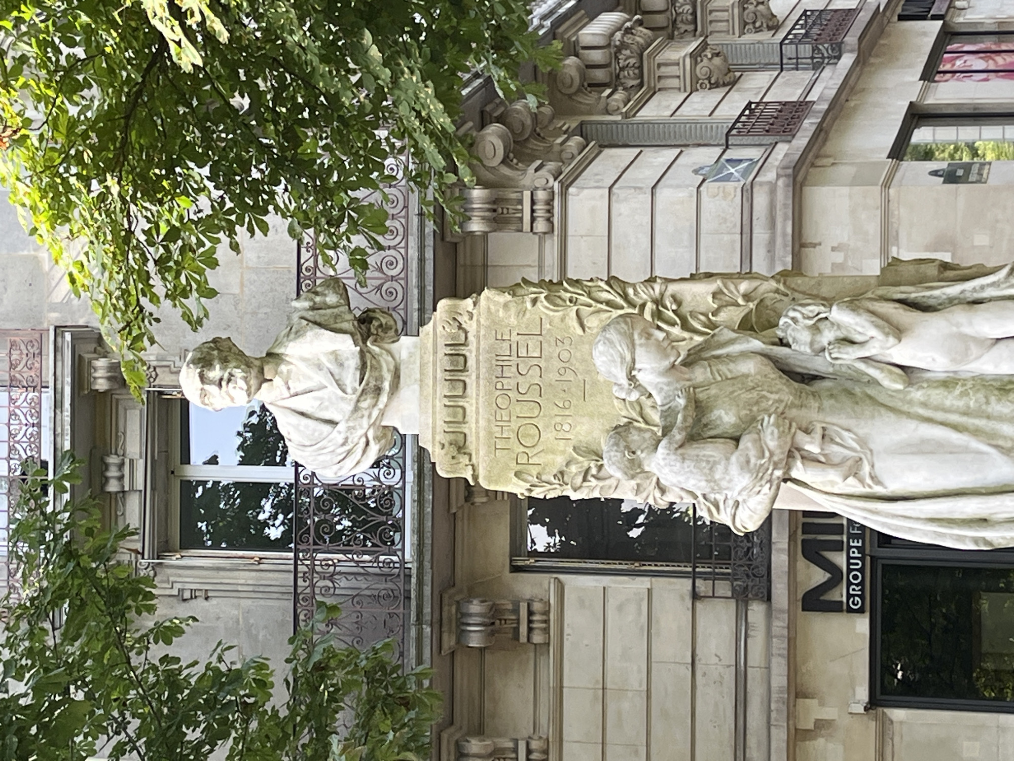 The image depicts a marble statue in Paris, France. The statue is a bust of a man with a beard, likely a historical figure, perched atop a larger pedestal. Below the bust, a sculpture features a mother embracing a child while another child looks upward. Inscriptions on the pedestal read "THEOPHILE ROUSSEL" and "1816-1903". The statue is situated in front of a building with a stone facade, ornate balconies, and large windows. Green foliage from nearby trees partially obscures the upper portion of the statue and the building. The lower part of the image shows a store with a sign that reads "MII GROUPE". The lighting suggests it is daytime with sunlight filtering through the leaves.