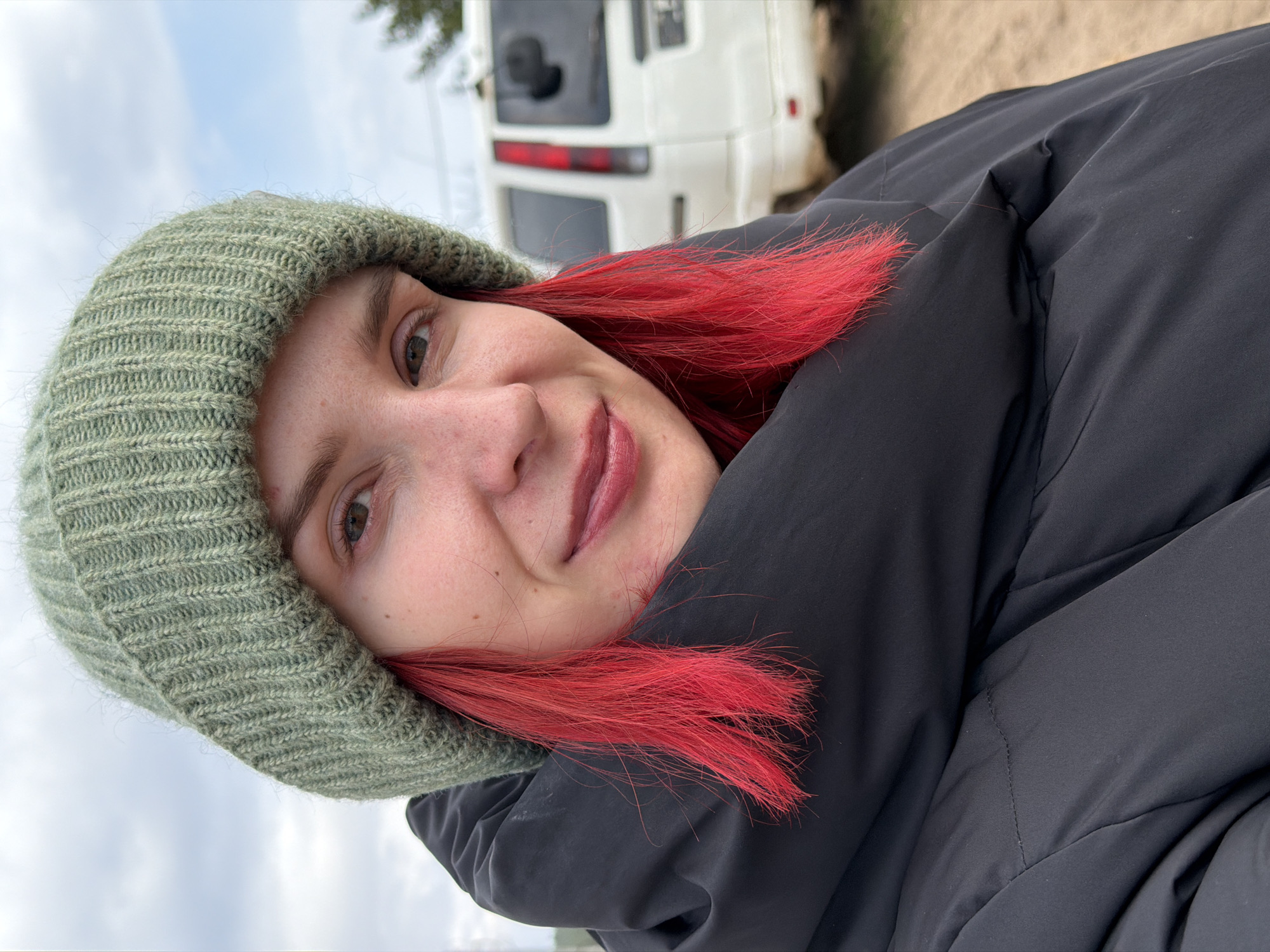 A young individual, likely female, is featured in a tilted selfie, looking directly at the camera with a friendly smile. She has fair skin, possibly with some freckles, and striking bright red hair that contrasts sharply with her attire. She is wearing a muted sage green, chunky knit beanie and a dark, possibly black, puffy winter jacket. Her lips are slightly parted in the smile, and her eyes appear to be brown or hazel.
The background is blurred, indicating a shallow depth of field. A white van or bus, possibly a camper or shuttle, with dark windows and red taillight accents, is visible behind her. Above the vehicle, a hint of a tree or foliage can be seen. The sky, visible in the upper left, is a light blue, suggesting it might be overcast or hazy. Some light-colored ground, possibly dirt or sand, is visible in the upper right background. The thick winter clothing suggests cool or cold weather, aligning with the provided location context of Chaikovsky, Russia. The overall scene depicts a casual, cheerful moment outdoors during daytime. No text is visible in the image.