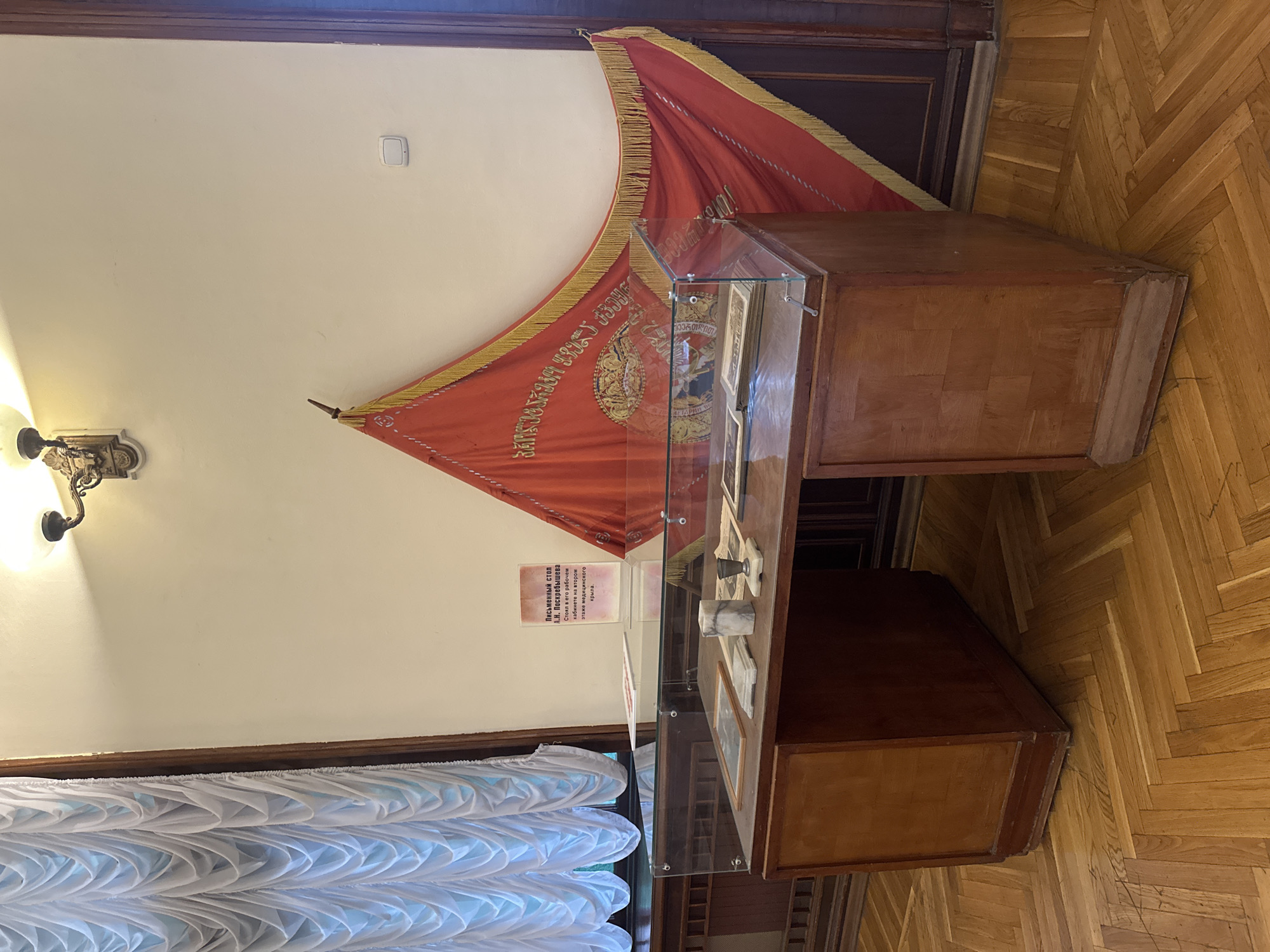 The image shows a room with a hardwood floor, a light switch on a white wall, and a wall sconce. There is a red banner with gold fringe hanging on the wall above a display case made of wood and glass. Inside the case are several items, including a small bell and framed pictures. A sign on the left side of the display case contains text in Russian. A white, decorative curtain is in the lower left corner. The sign mentions a written table of A.N. Poskrebysheva, and that it stood in his workroom on the second floor of the medical wing, meaning this picture was taken in a museum located in Sochi, Russia.