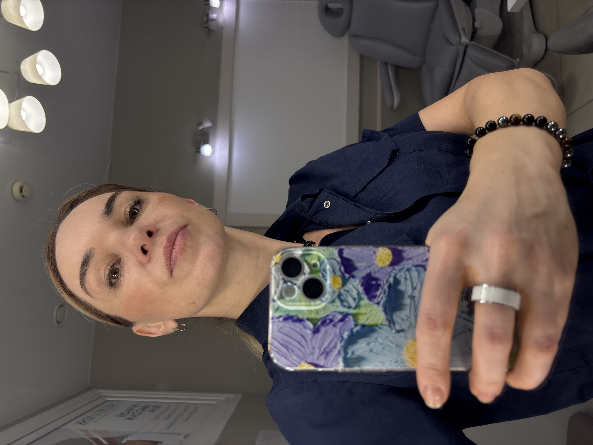 The image shows a woman taking a selfie with a floral-patterned phone case. She is wearing a dark blue collared shirt and jewelry including a bracelet and a ring. The background includes a light fixture, a wall, and what appears to be a dental or medical chair. While not definitively confirmed, the presence of a dental chair suggests the location is likely a dental or medical office. The location in Murino, Russia, is not verifiable from the provided image.