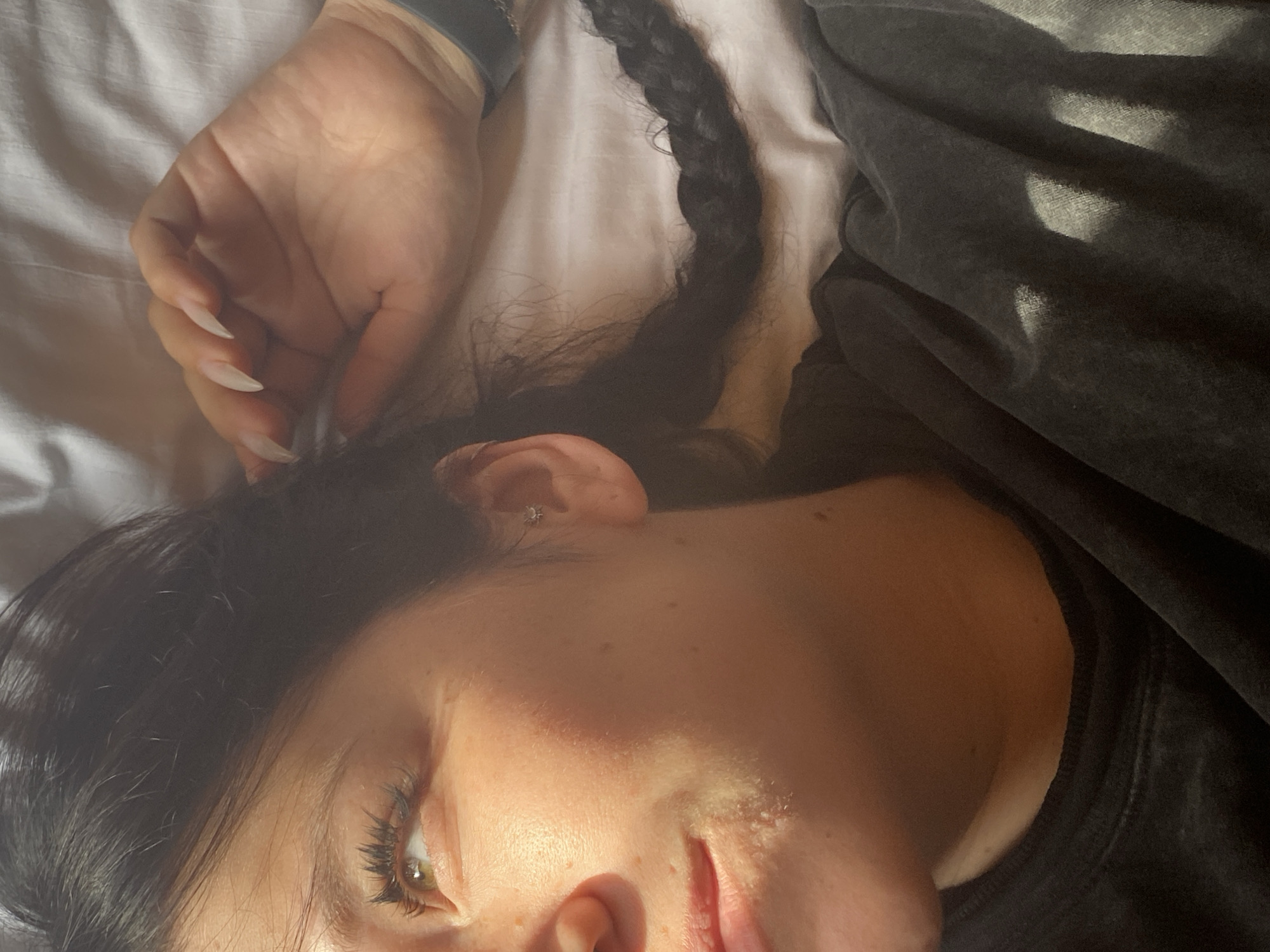 The image shows a close-up of a person's face and upper body. The person has dark hair, some of which is braided and rests on a white surface, possibly a bed. Sunlight is hitting the person's face, creating highlights and shadows. The person is wearing a dark, possibly grey, sweatshirt. A hand with long, light-colored fingernails is resting near the person's head. There is a small, star-shaped earring in the person's ear. The background appears to be a white sheet or pillowcase. The image does not contain any specific landmarks or context that would indicate a location in Krasnoflotskoe, Russia.