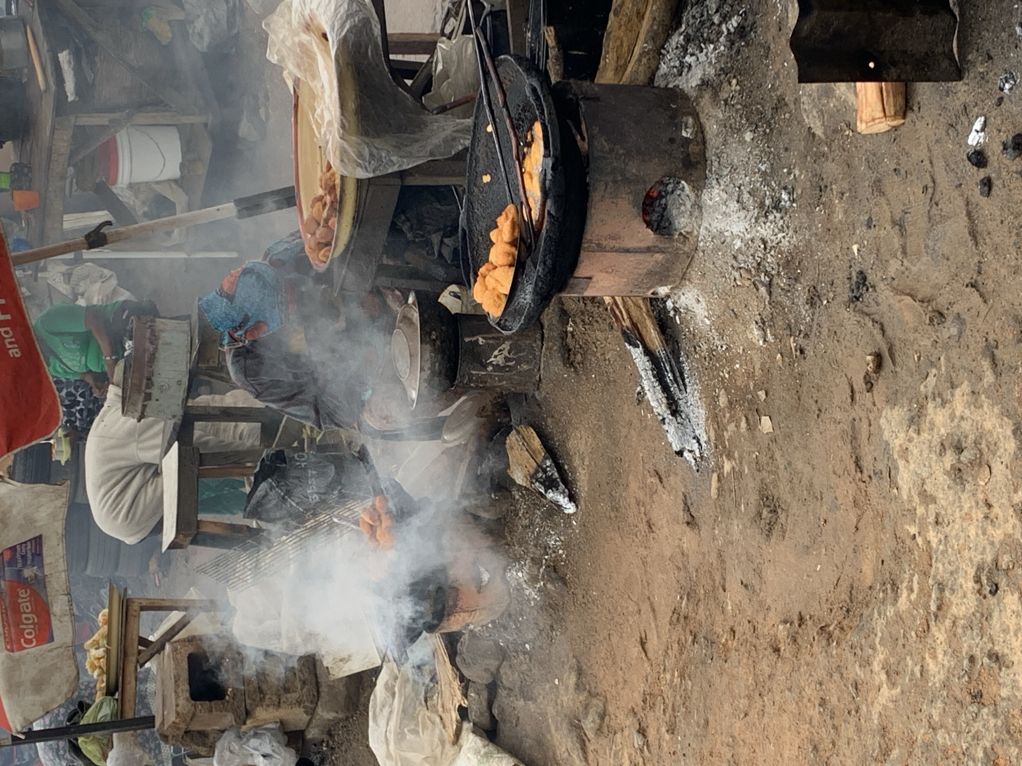 The image depicts a street food vendor operation in Rafin Par, Nigeria. A metal pot sits on a wood-burning stove, frying what appears to be puff-puff (a fried dough). Smoke billows from the stove, obscuring the surrounding area. To the left, a large bowl of uncooked dough sits on a makeshift table. People are visible in the background, partially obscured by the smoke. A sack of "Colgate" tooth paste products sits off to the side. The ground is dirt and scattered with ash and debris.